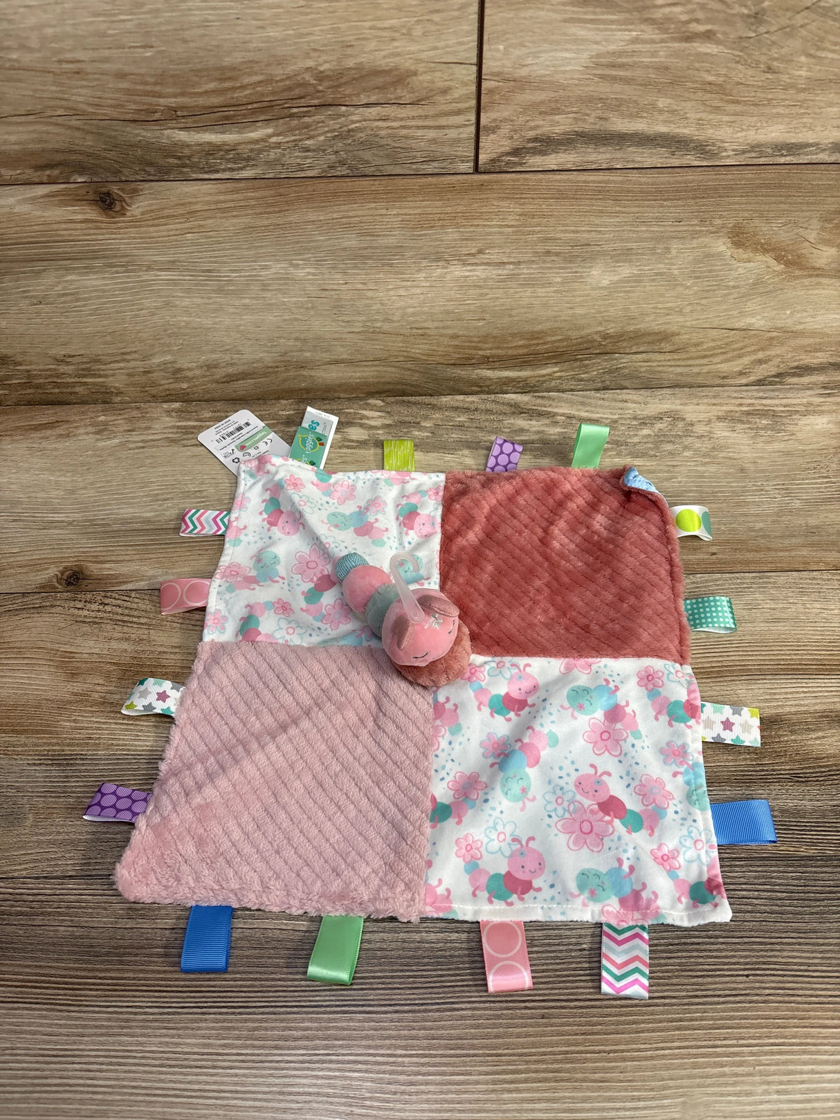 Soft baby sensory tag blanket with pink and floral patches and a small caterpillar plush on wooden floor