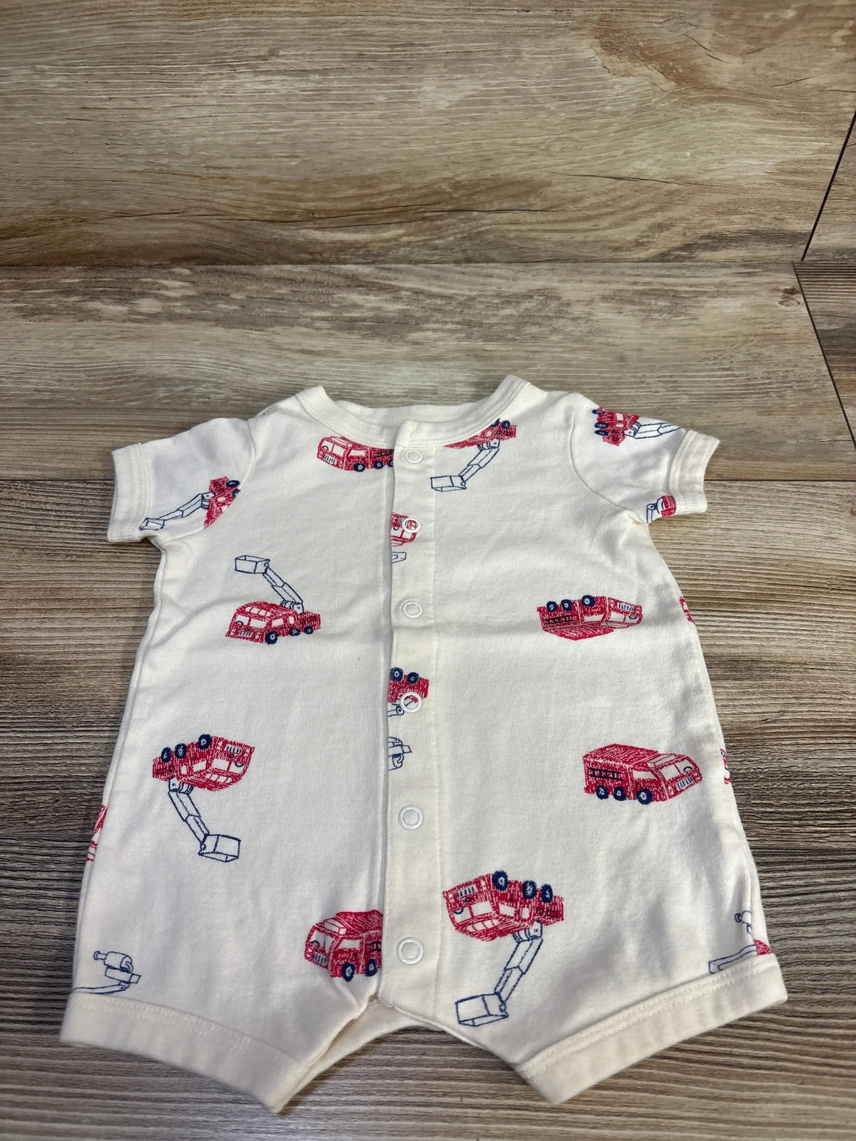 Front flat lay of Carter’s button-front shortie romper showing red truck print and snap buttons on light wood background