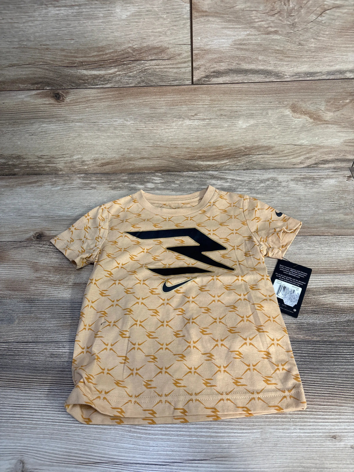 Front view of Nike x 3BRAND toddler shirt in sesame beige with lightning pattern and black swoosh on wood flooring