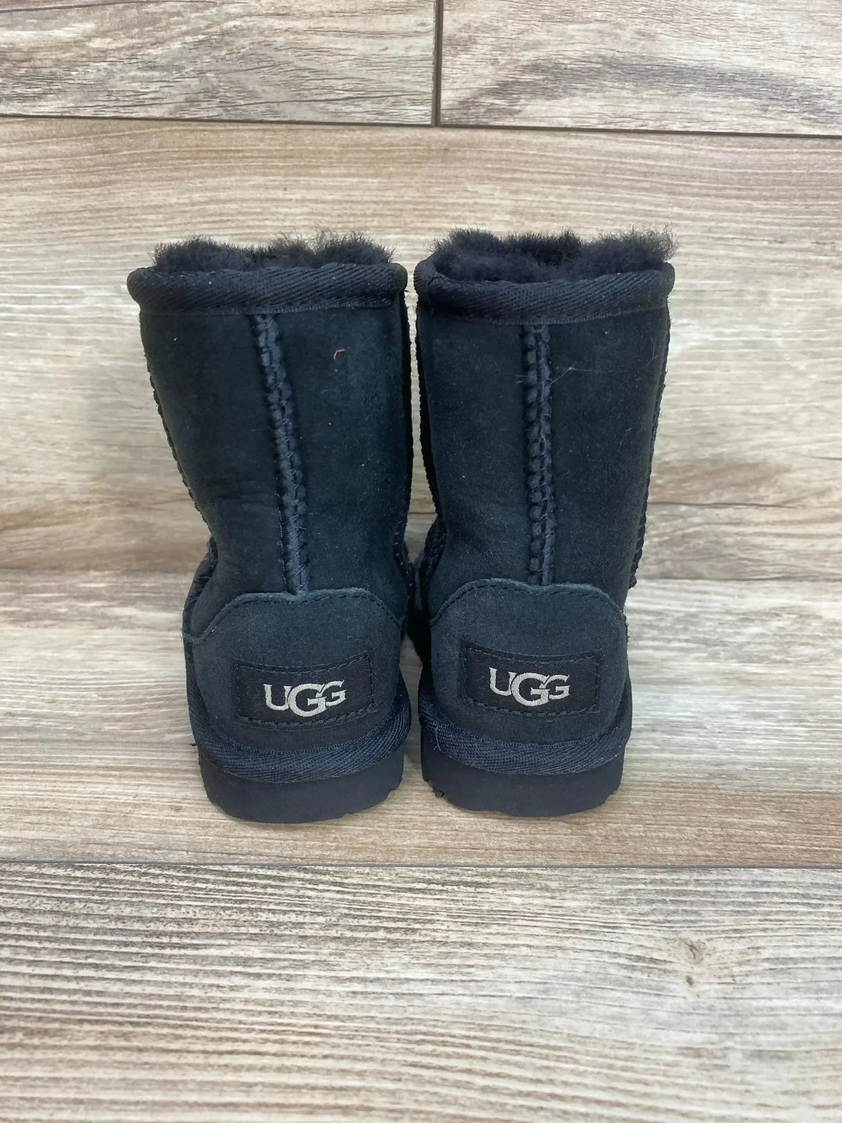 Rear view of UGG toddler Classic II boot in black showcasing suede exterior, plush sheepskin lining and UGG heel logo on wood floor background