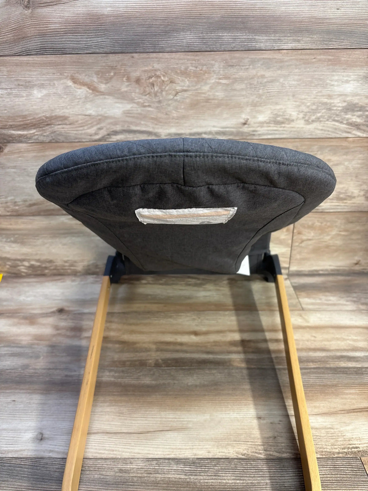 Rear view of ANGELBLISS portable baby bouncer in grey showing padded backrest, wooden rocker legs, and rear carry handle on wood-plank background