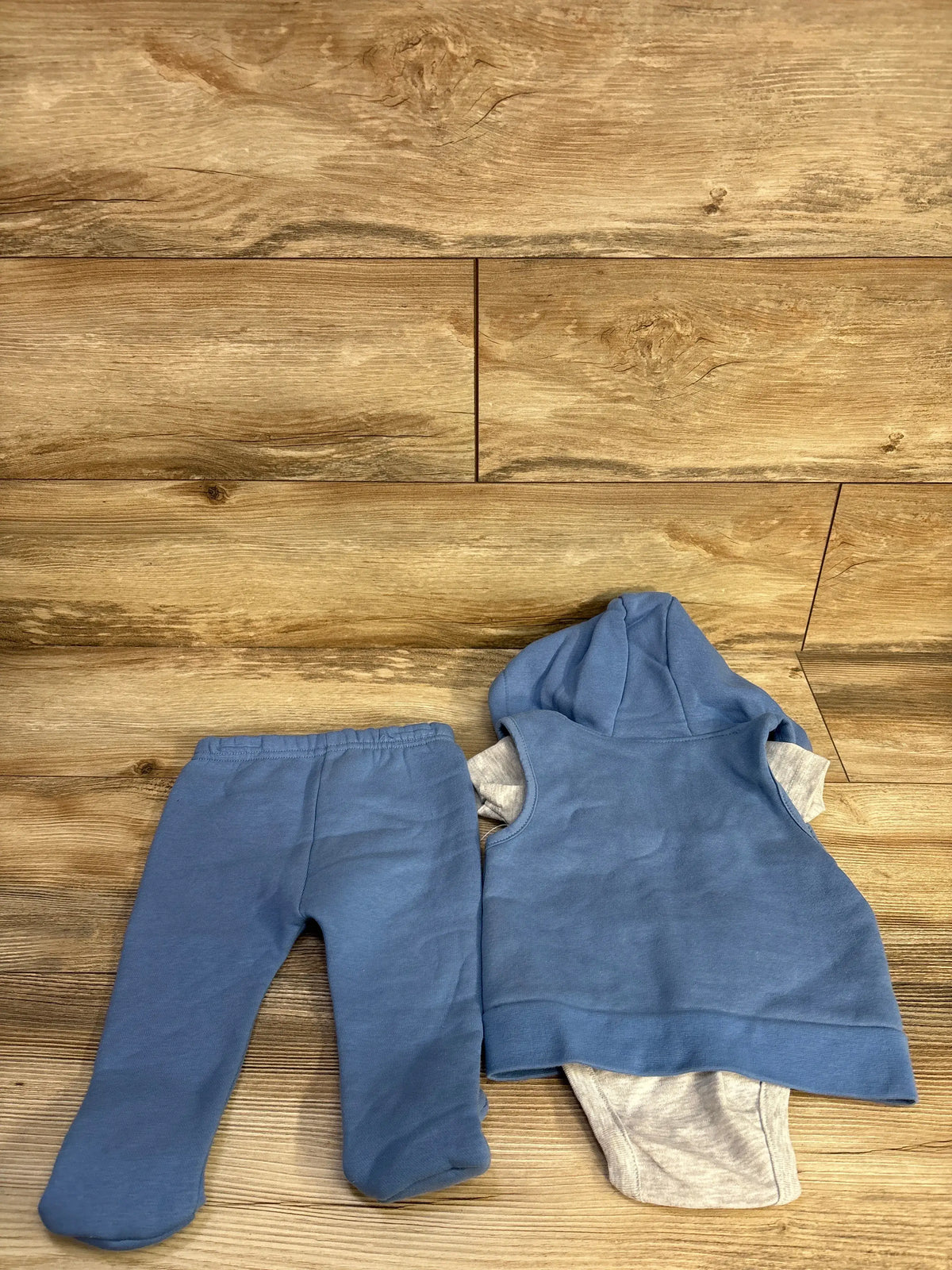 Top view of blue DKNY three-piece baby set showcasing hooded vest, bodysuit, and footed pants on a wood floor