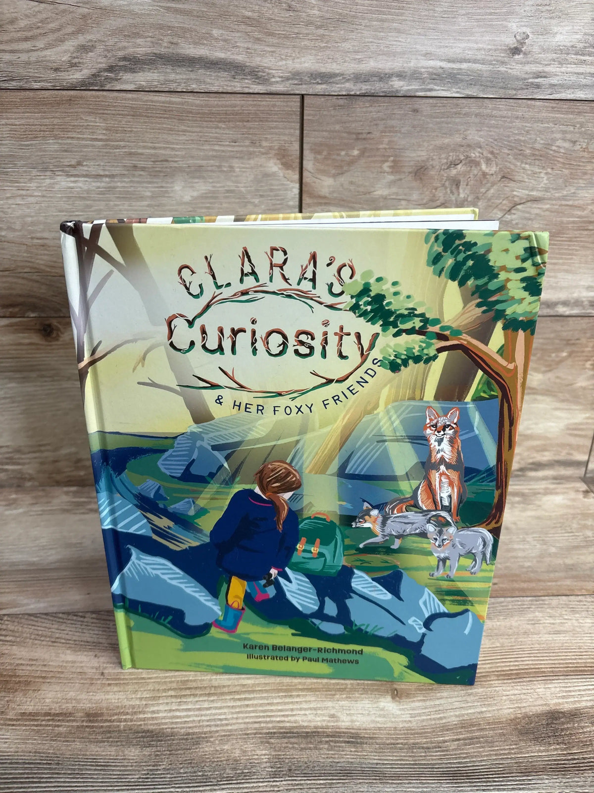 Front view of Clara's Curiosity and Her Foxy Friends hardcover showing cover illustration of a girl and three foxes on wood