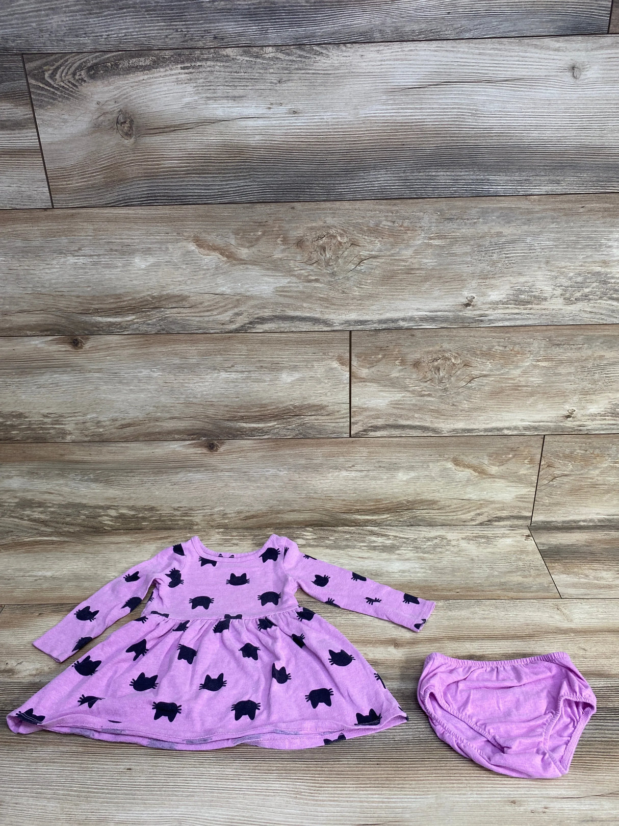 Flat lay of purple long sleeve baby dress with black cat print and matching solid diaper cover on wood plank floor