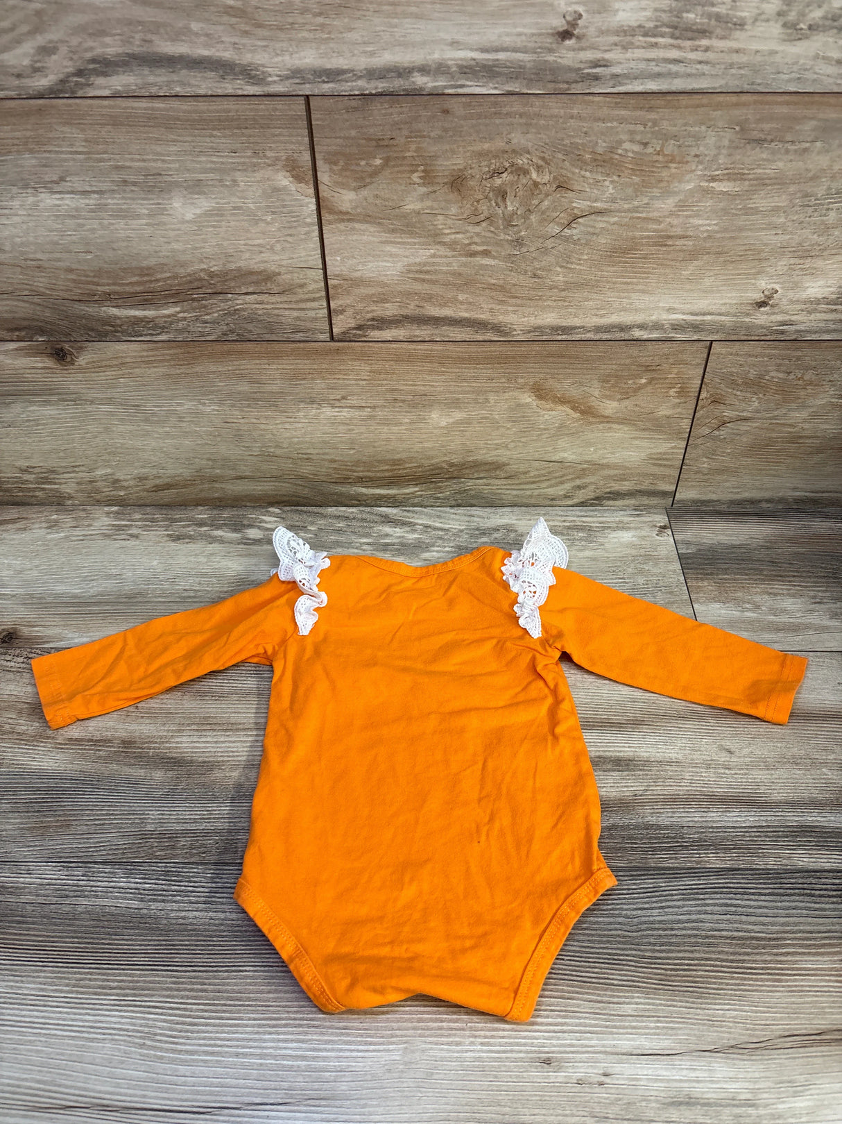 Flat lay of orange long-sleeve baby bodysuit showcasing white lace ruffle shoulders on wood background