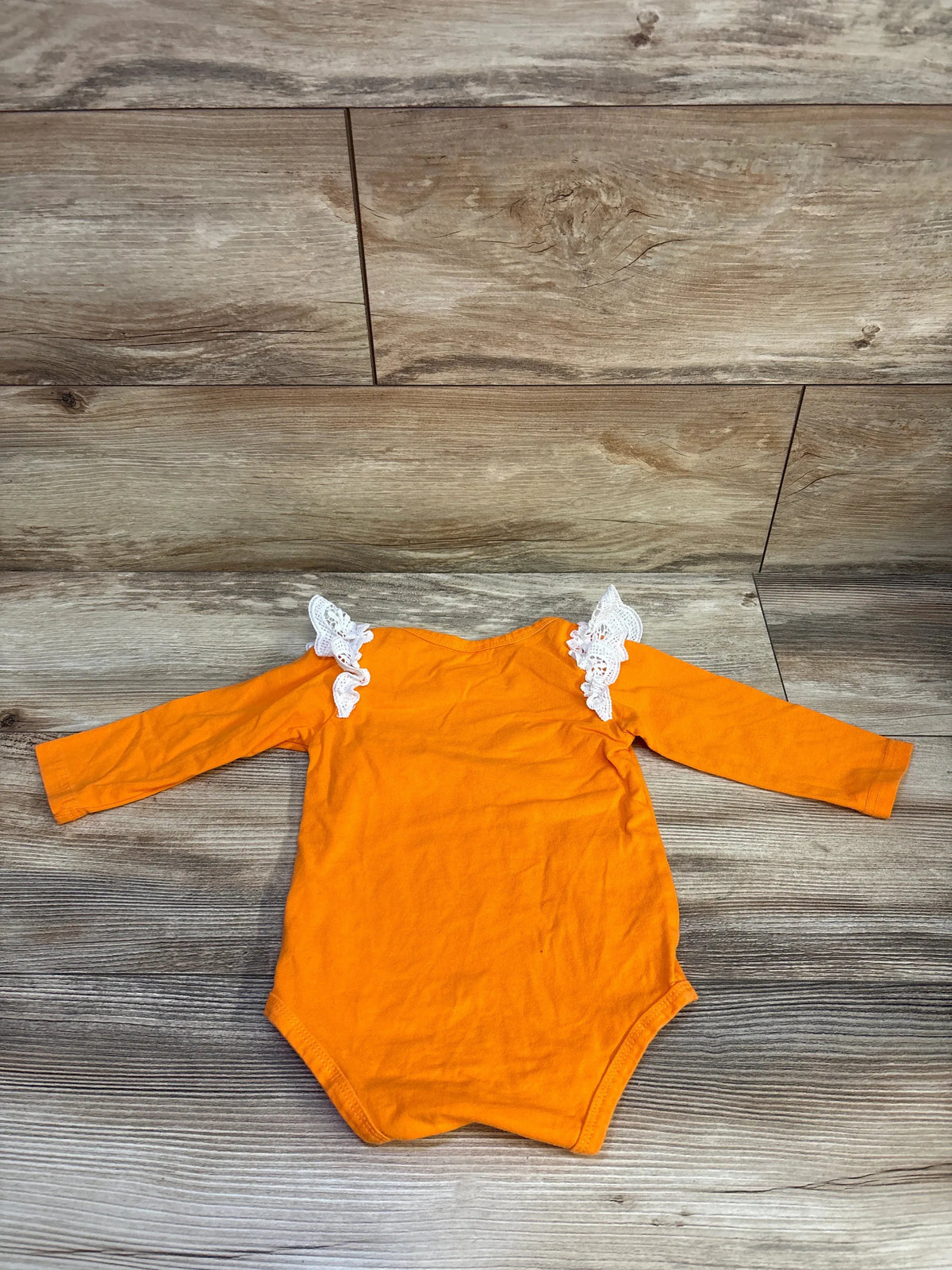 Flat lay of orange long-sleeve baby bodysuit showcasing white lace ruffle shoulders on wood background
