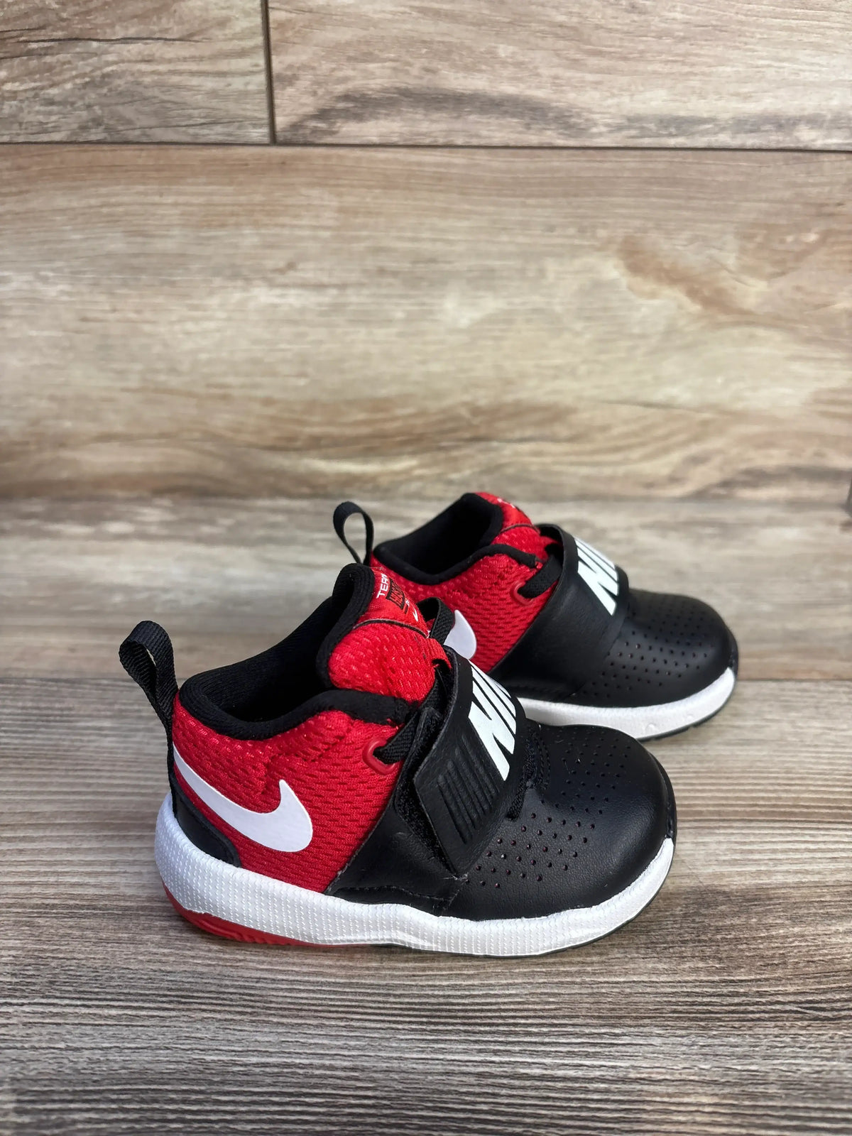 Front three-quarter view of Nike Team Hustle D8 black and university red sneakers showing perforated toes on a wood floor