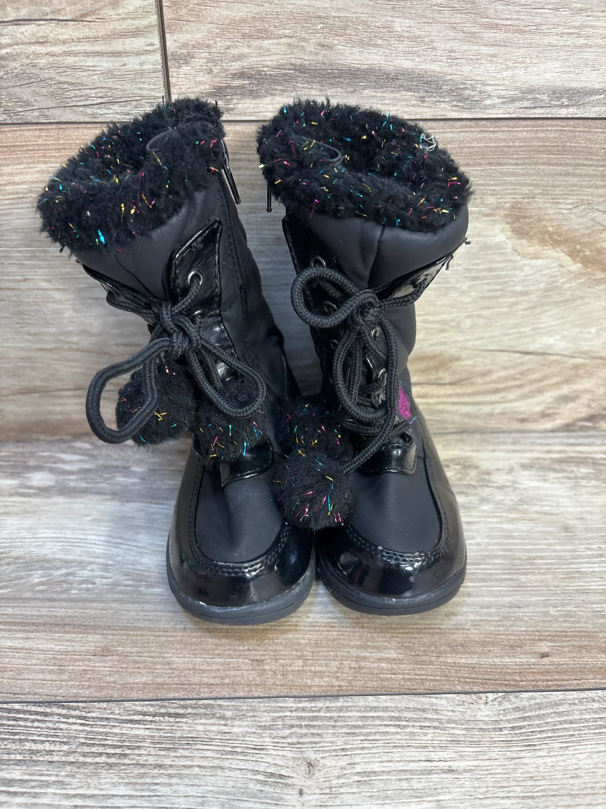 Black toddler winter boots with faux fur trim, glitter accents, and pom pom laces on wood floor
