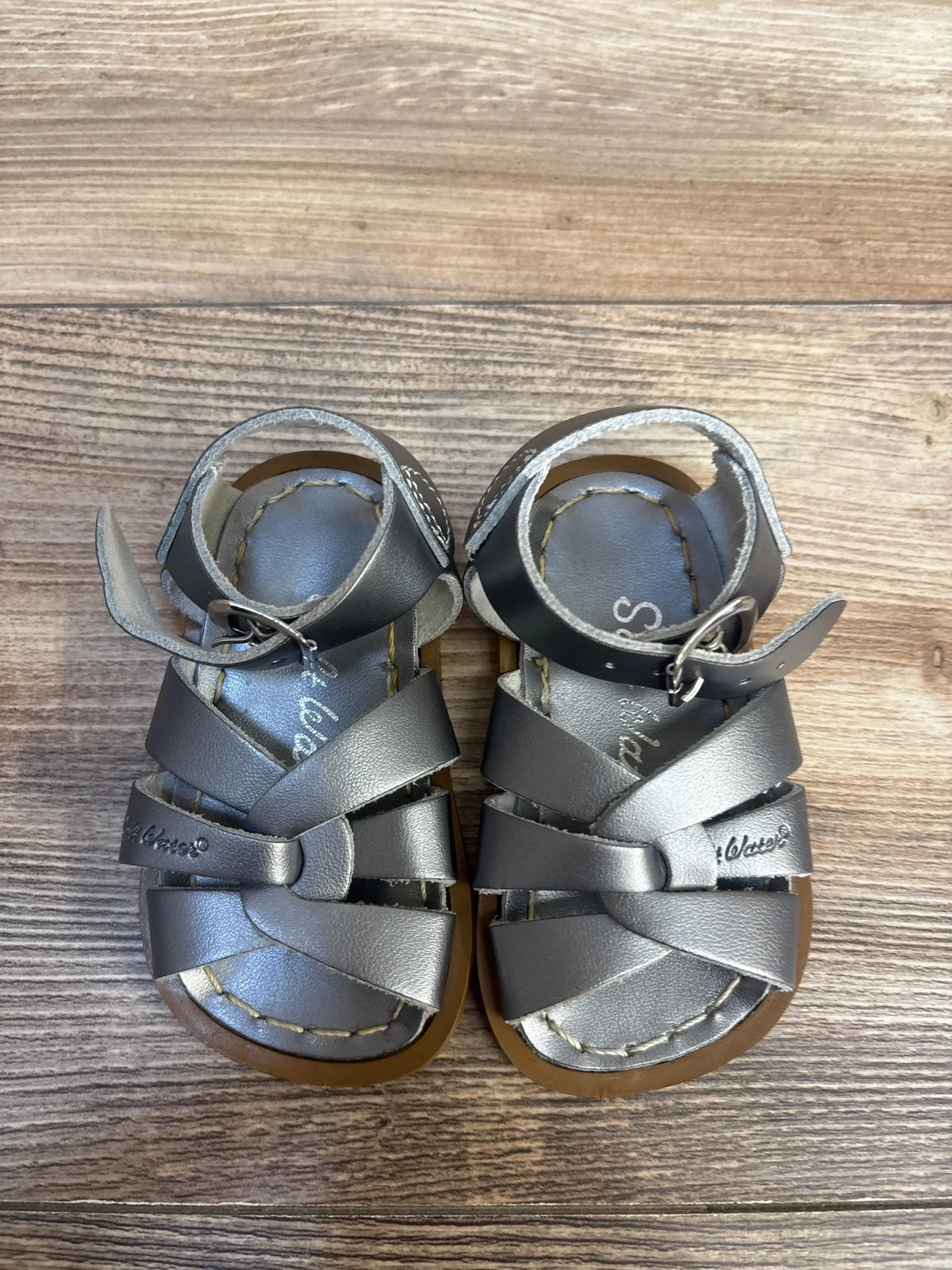 Top-down of Salt-Water Original toddler sandals size 3C showing silver crisscross straps, ankle buckle and brown stitched soles on wood