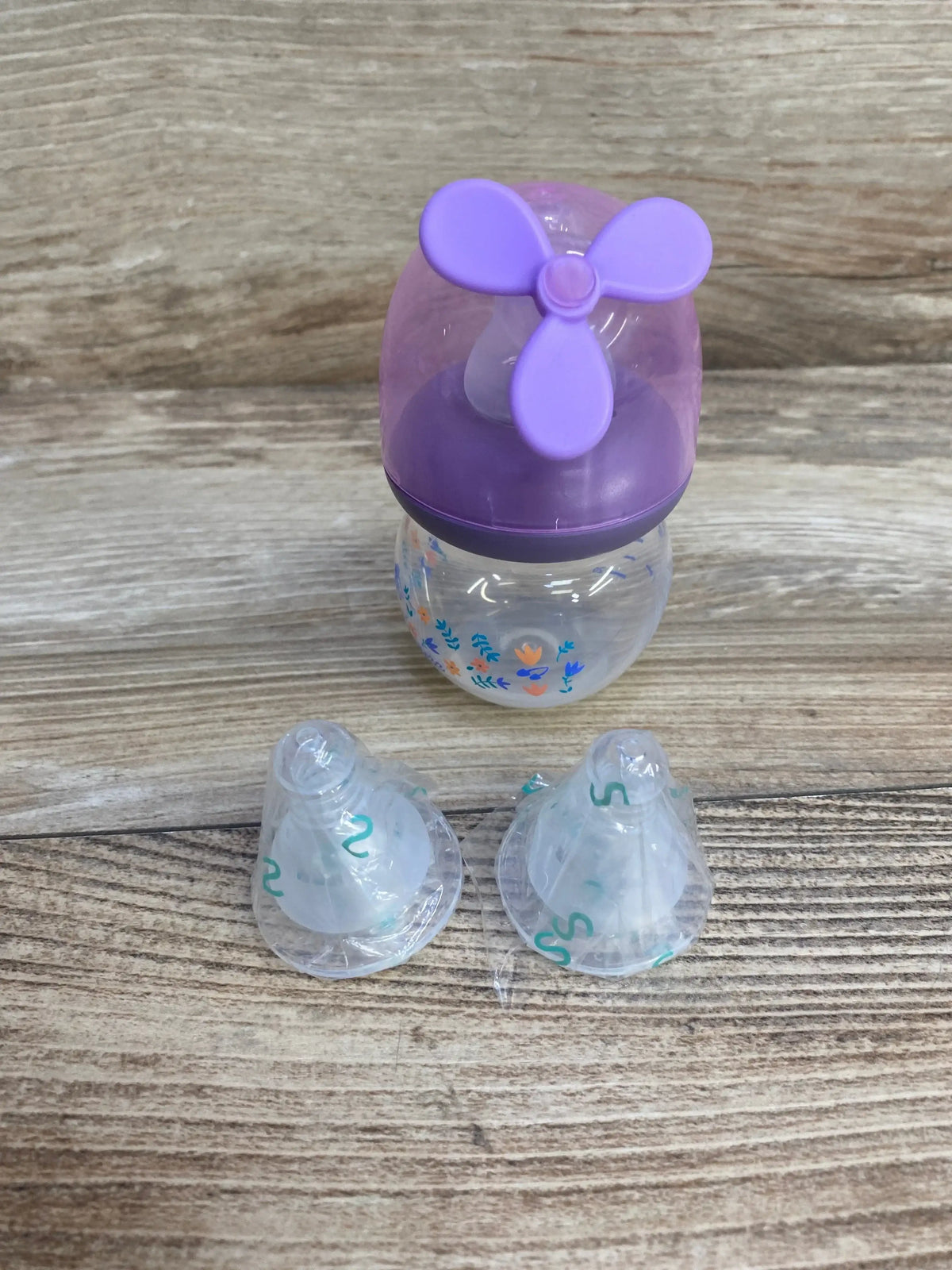 Front view of MINITree 2 oz purple baby bottle with floral print, purple propeller-style cap and two extra sealed nipples on a wood surface
