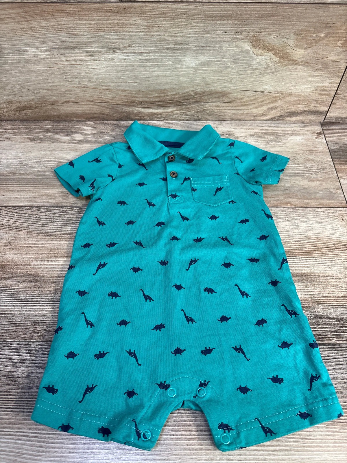 Flat lay of green Simple Joys dinosaur polo shortie romper showcasing print, collar, pocket, and snaps on wood