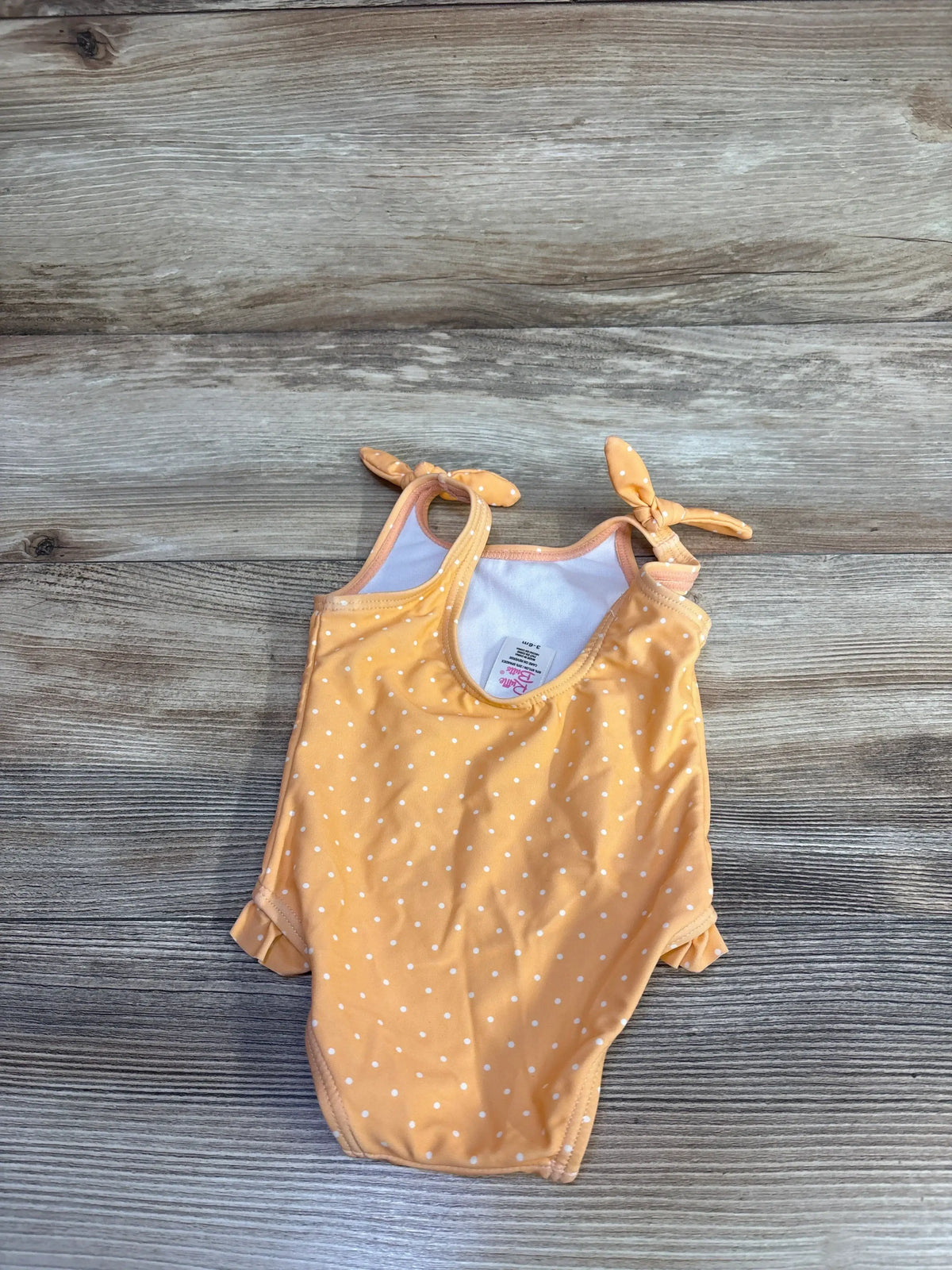 Front view of Ruffle Butts melon polka dot one-piece baby swimsuit showing tied shoulder straps and ruffle leg trim on wood background