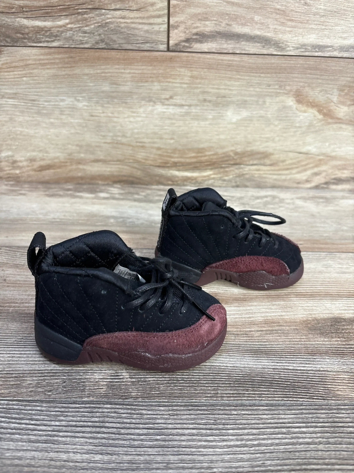 Angled side view of A Ma Maniére x Air Jordan 12 toddler sneaker (5C) with black upper and burgundy suede on wood floor