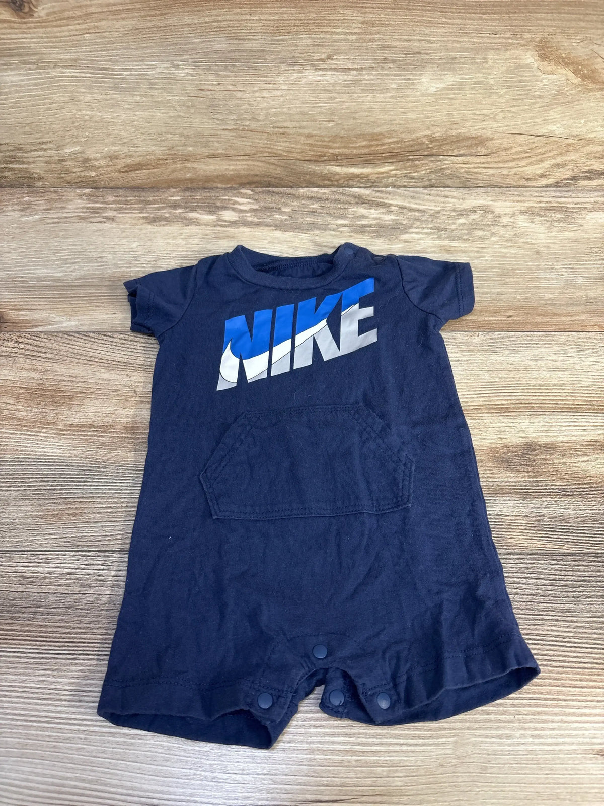 Flat lay of navy baby shortie romper with blue and white NIKE logo, front kangaroo pocket and snap crotch on wood background