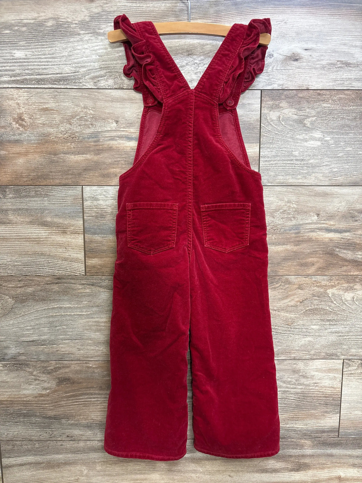 Back view of Baby Gap red velvet heart overalls showcasing ruffle straps and two back patch pockets on a wooden hanger