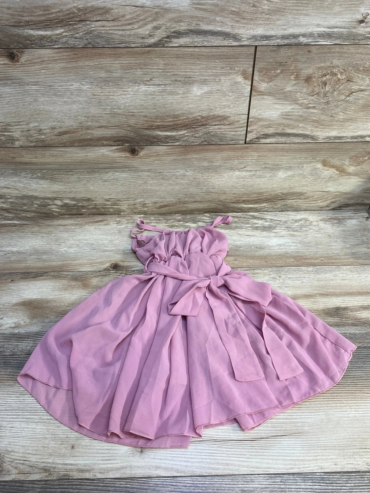 Flat lay of pink sleeveless baby dress (size 3-6 months) showing ruched bodice, tie straps and sash bow on wood floor