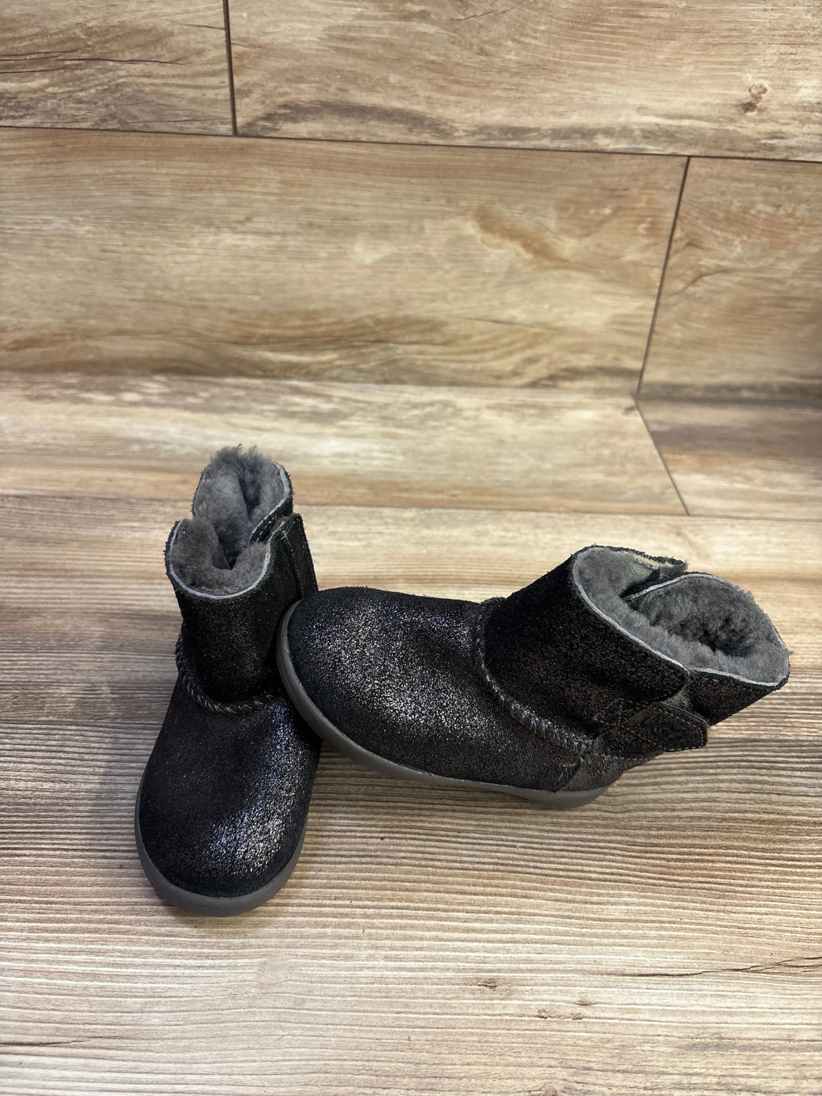 Angled view of black UGG Keelan metallic glitter boots showcasing gray fuzzy lining on a wood-look floor