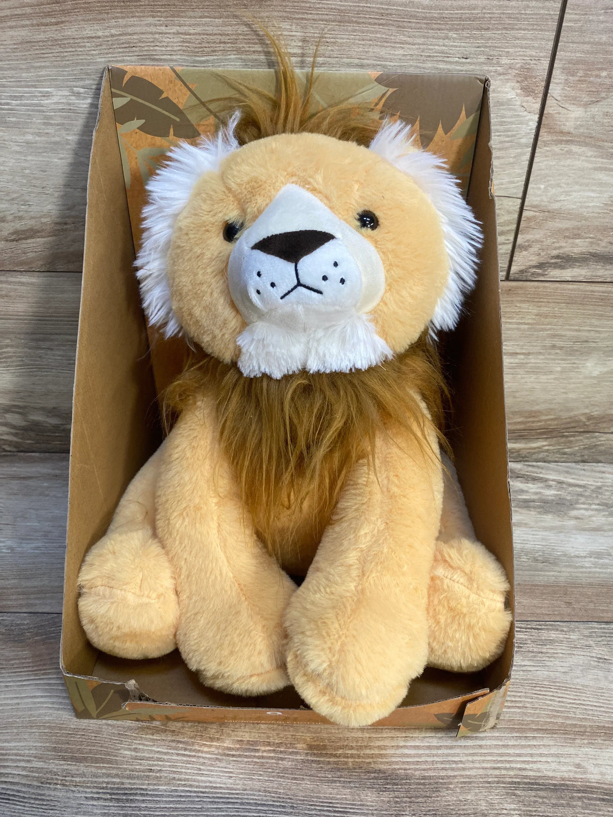 Front view of Aroma Home Hot Hugs microwavable plush lion toy showing white facial fur and brown mane seated in its cardboard box on a wood floor