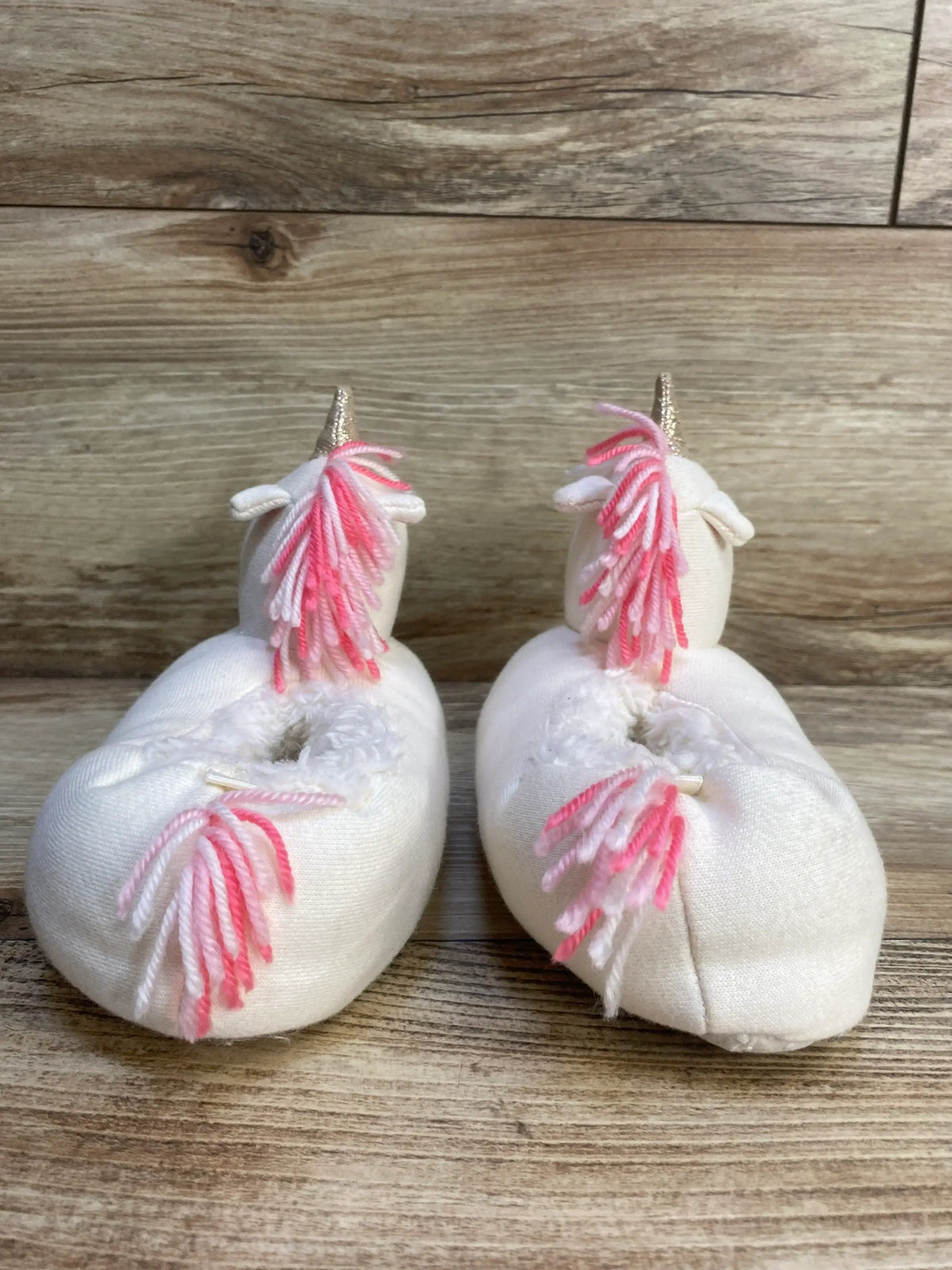 Plush white baby unicorn slippers with pink yarn manes on wood background