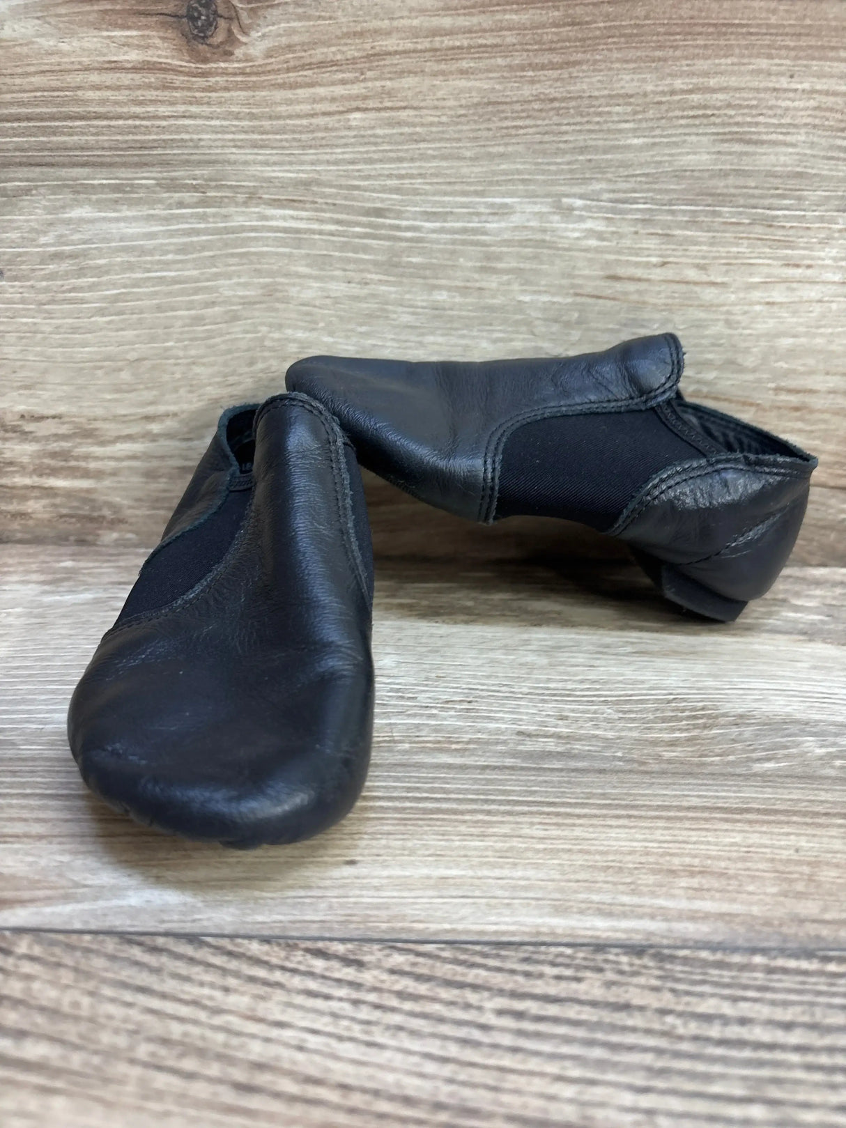 Front three-quarter view of Capezio EJ2C black leather jazz shoes showing elastic side panels and split-sole on wood floor