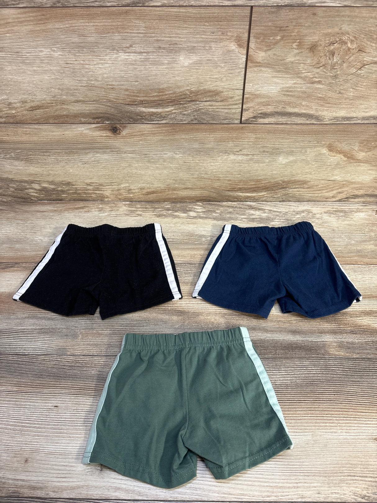 Flat lay of three Garanimals toddler pull-on shorts, black navy and olive, showing elastic waists and white side stripes on wood
