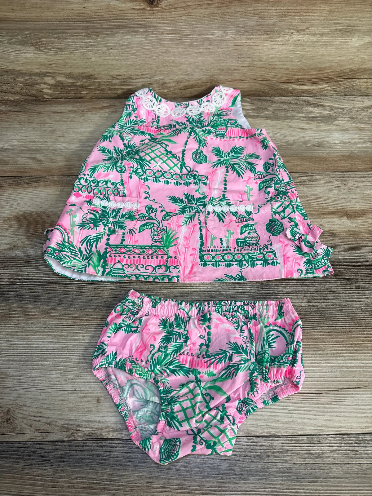 Pink and green tropical baby dress and bloomers set with lace trim on a wood background.