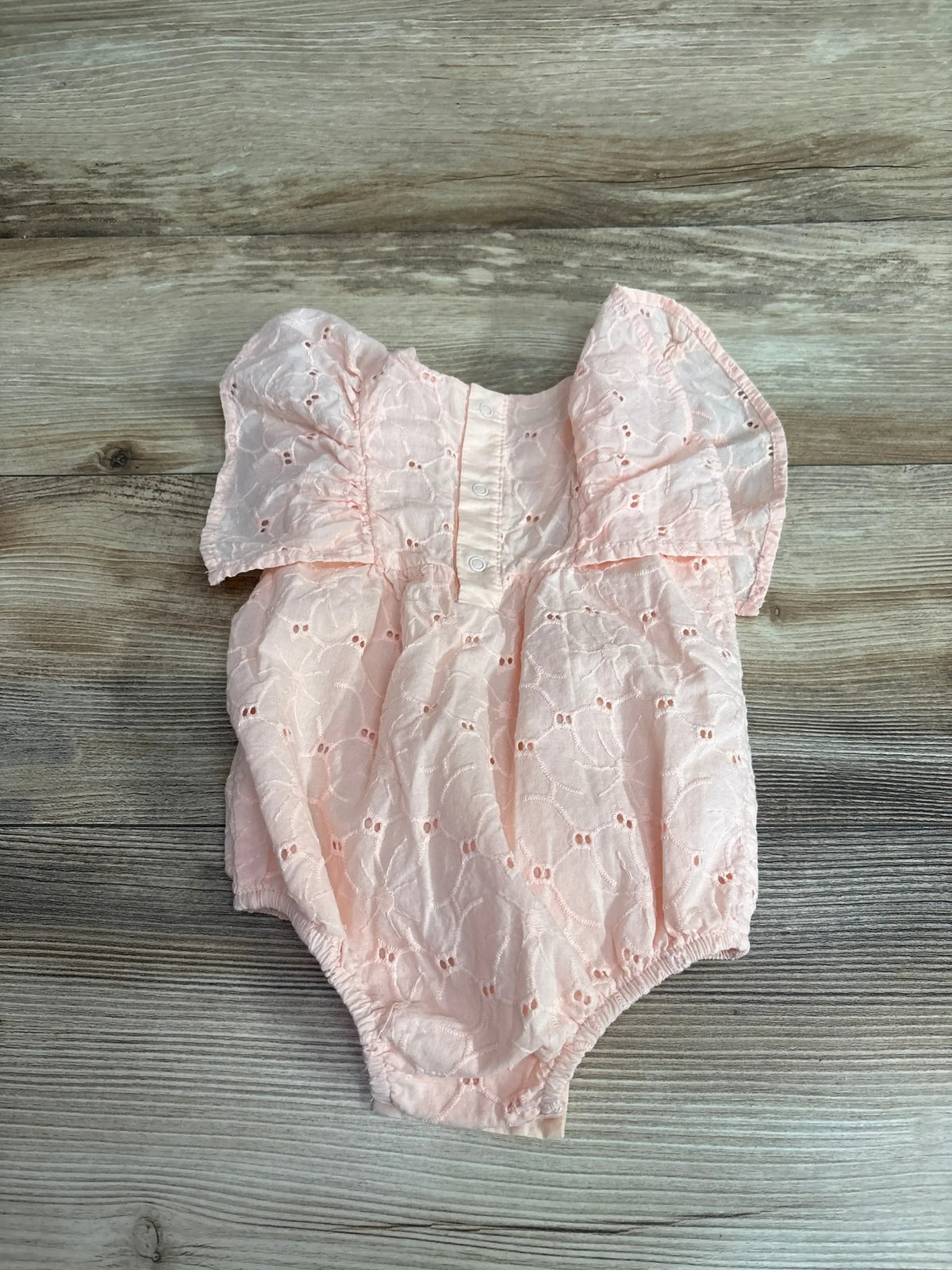 Front view of Cat & Jack blush eyelet bubble romper showcasing puff sleeves and button placket on a wood-grain background