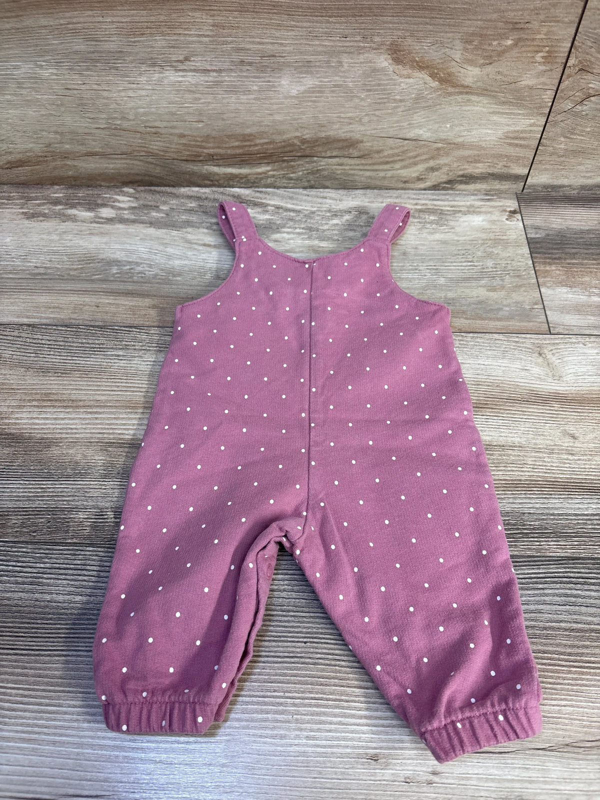 Front flat lay of pink polka dot baby overalls size 3M showing shoulder straps and elasticized ankles on wood floor