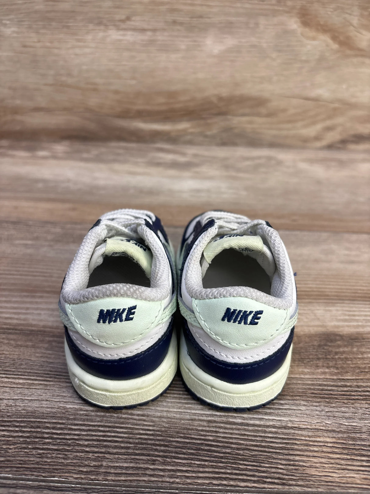Rear view of Nike Dunk Low TD sneakers showing NIKE heel tabs in midnight navy and volt tint on wood floor
