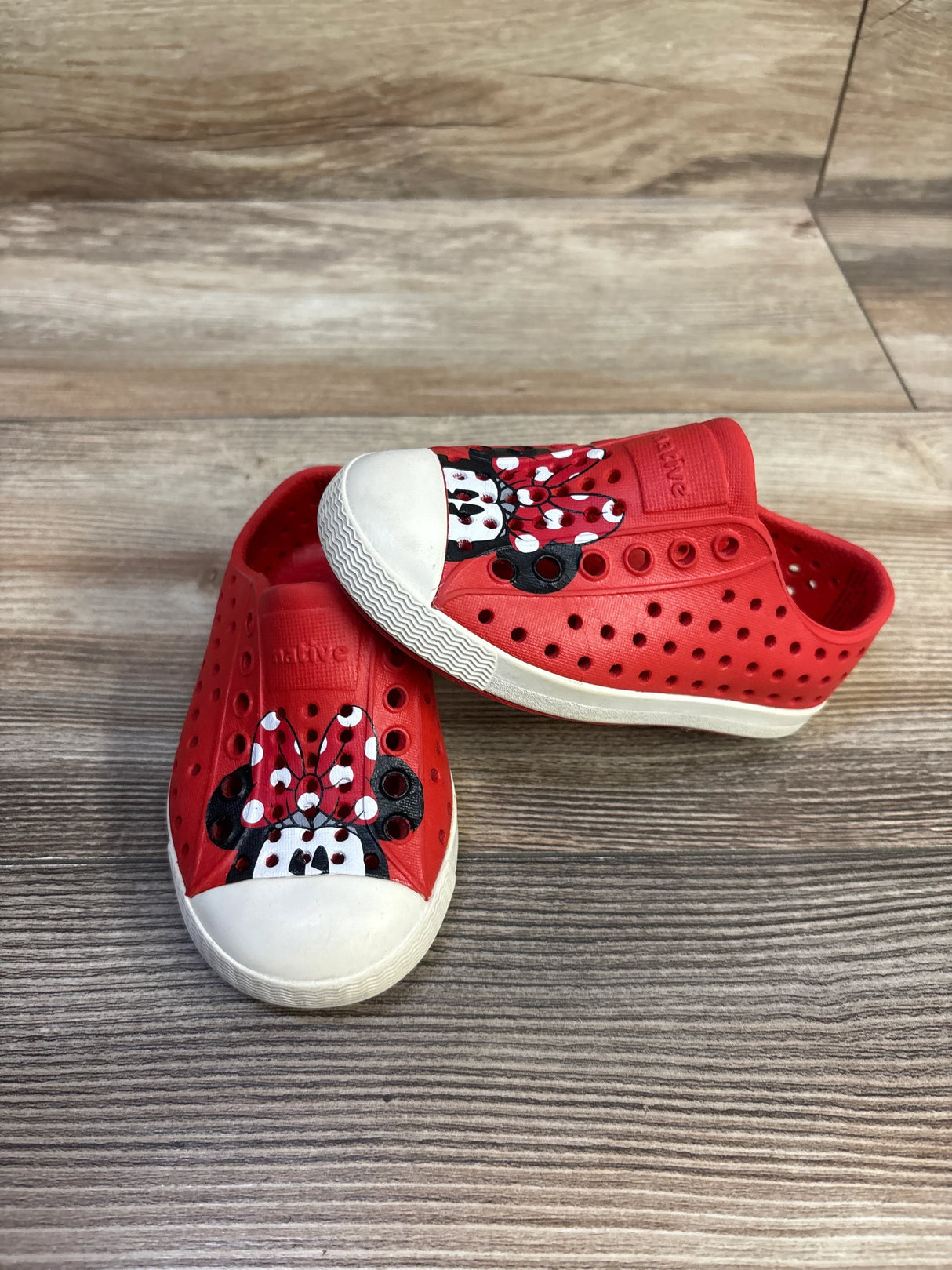 Angled front view of red Disney x Native Jefferson slip-on toddler shoe showing Minnie Mouse bow print on the white toe cap on wood background