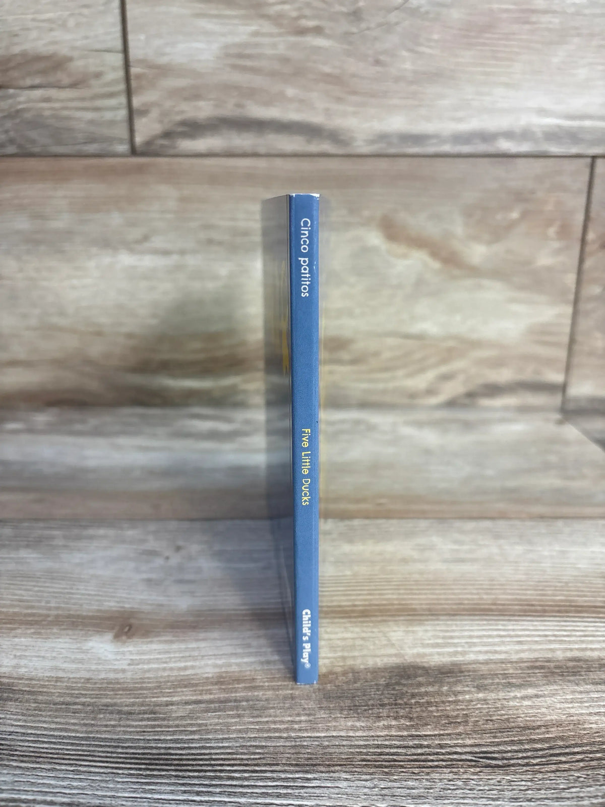 Spine view of Cinco Patitos and Five Little Ducks board book showcasing its blue bilingual spine on a wood grain background