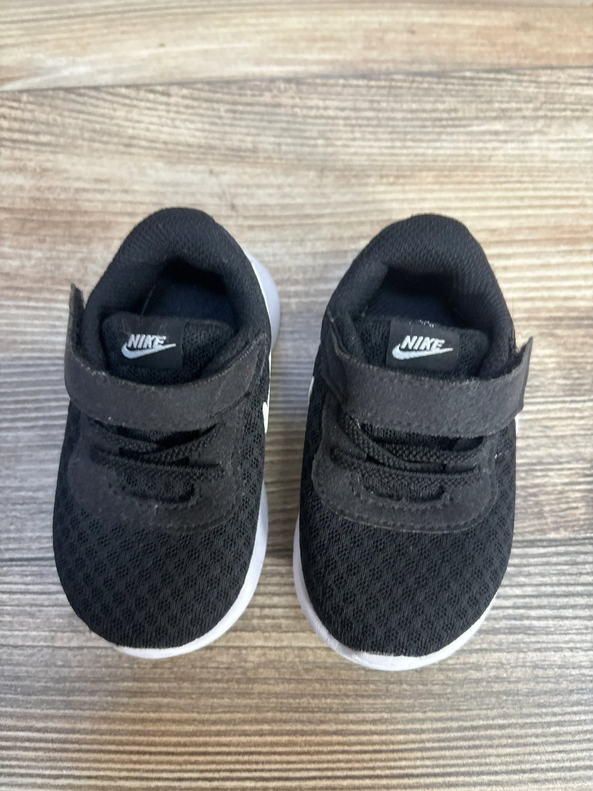 Overhead view of Nike Tanjun TDV toddler sneakers in black showing hook-and-loop straps and white soles on wood floor