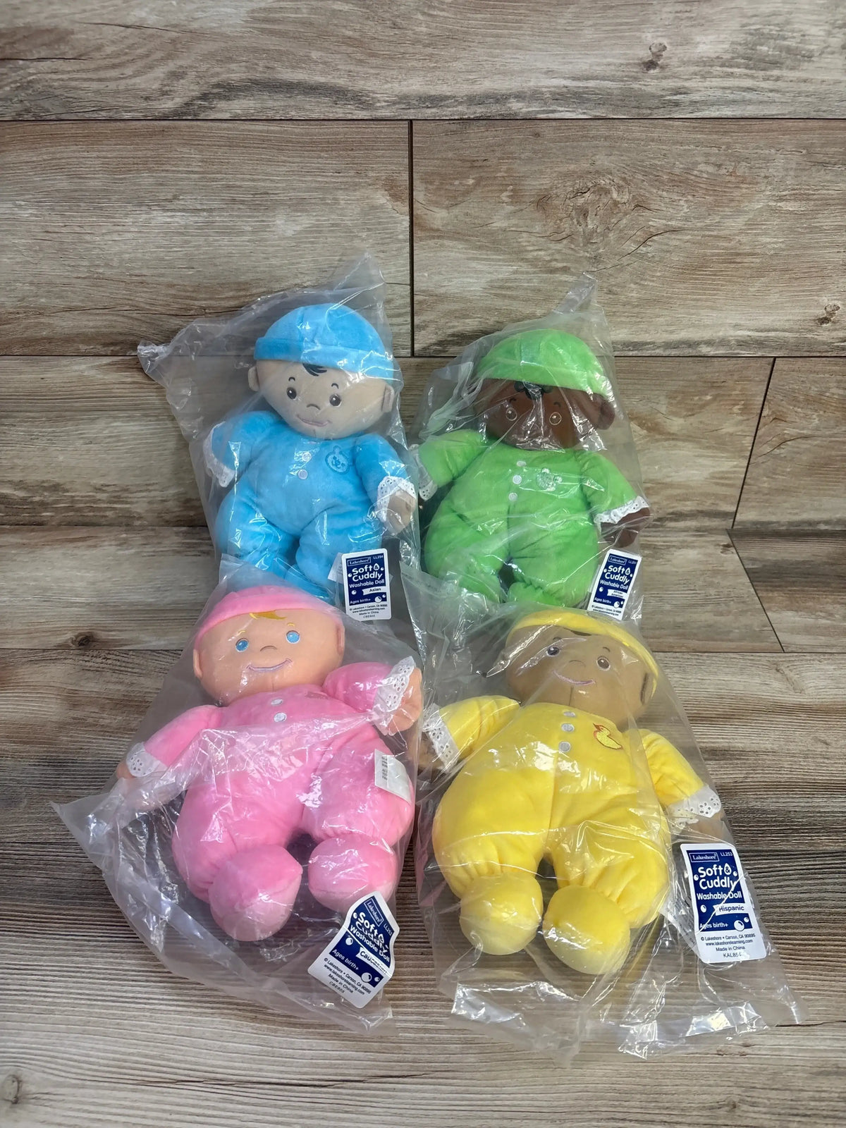 Front of four Lakeshore cuddle dolls in blue, green, pink and yellow with embroidered faces, lace cuffs on wood background