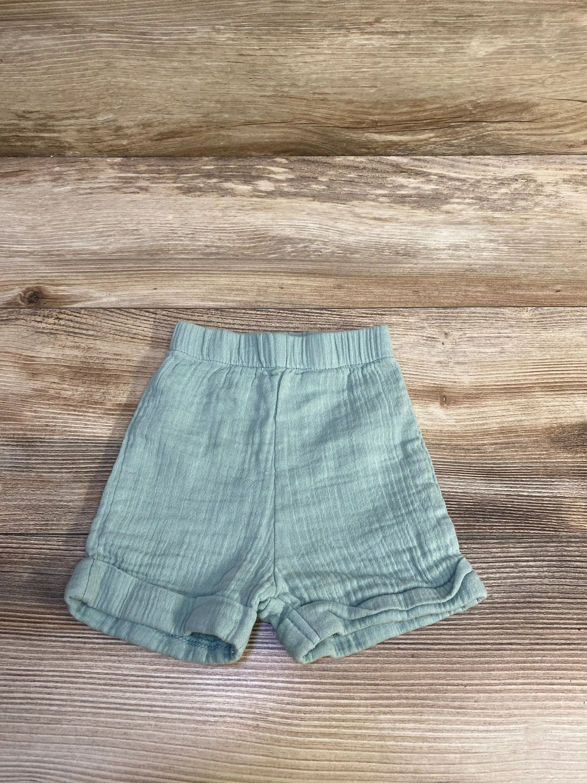 Front view of Embrace green gauze pull-on shorts, size 6 months, showcasing elastic waistband and rolled cuffs on a wood-grain background