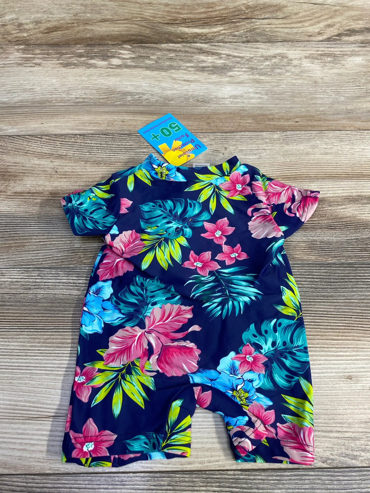 Flat lay of navy floral one-piece baby swimsuit 3M with short sleeves, attached shorts and SPF 50+ tag on wood background