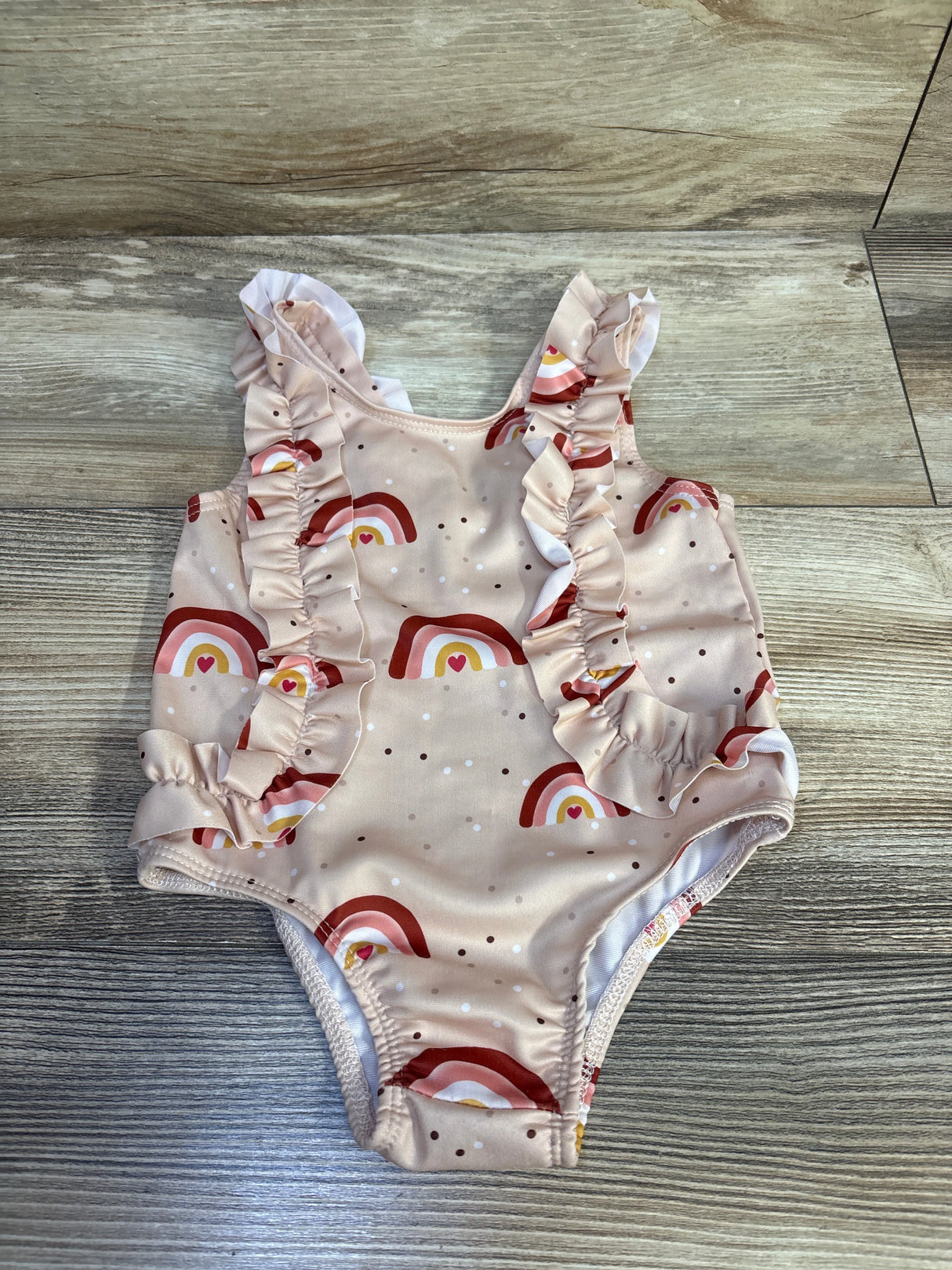 Front view of beige baby one-piece swimsuit with red and pink rainbow print and ruffled straps, laid flat on wood background