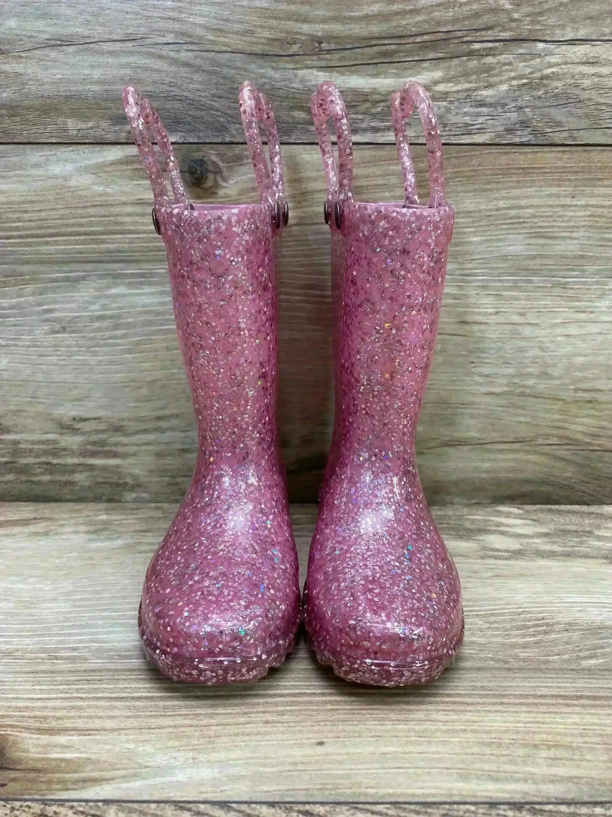 Front view of Western Chief toddler rain boots showcasing pink glitter finish and pull handles with wood plank background