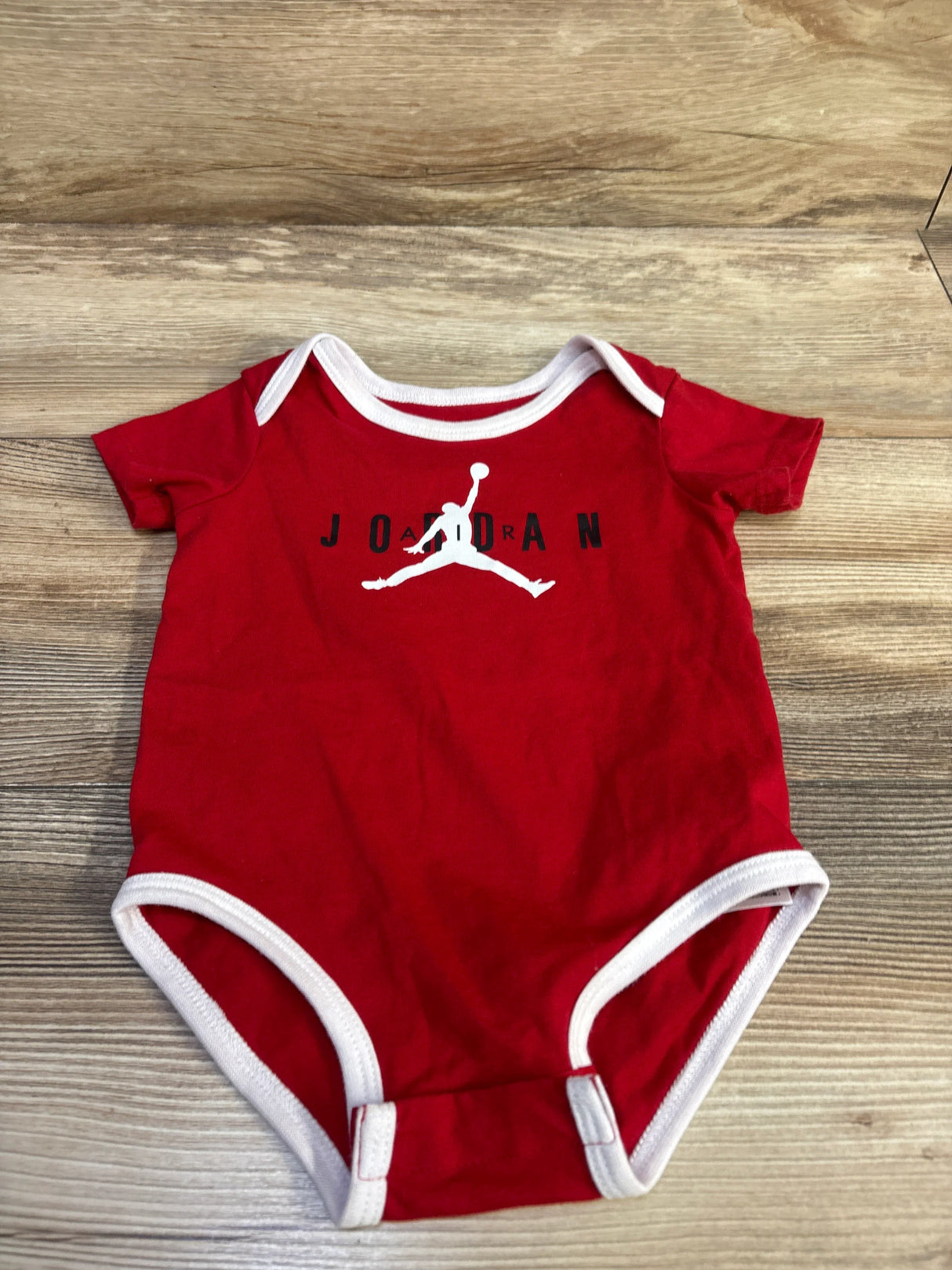 Top-down view of red Jordan baby bodysuit size 6-12 months showcasing white trim and Jumpman logo on light wood background