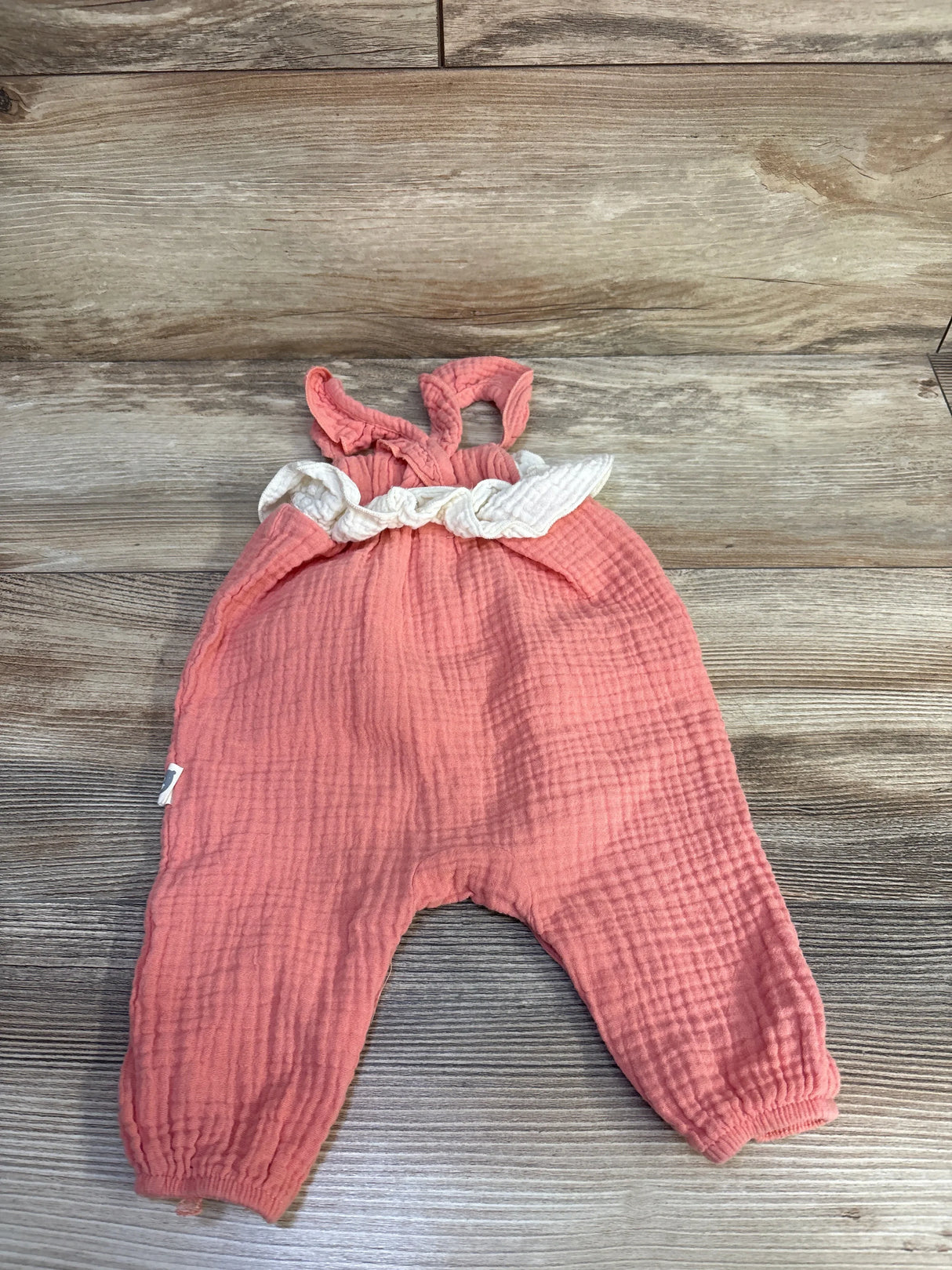Flat lay of pink gauze baby jumpsuit with white ruffle waist, tied shoulder straps and elasticized ankles on wood background