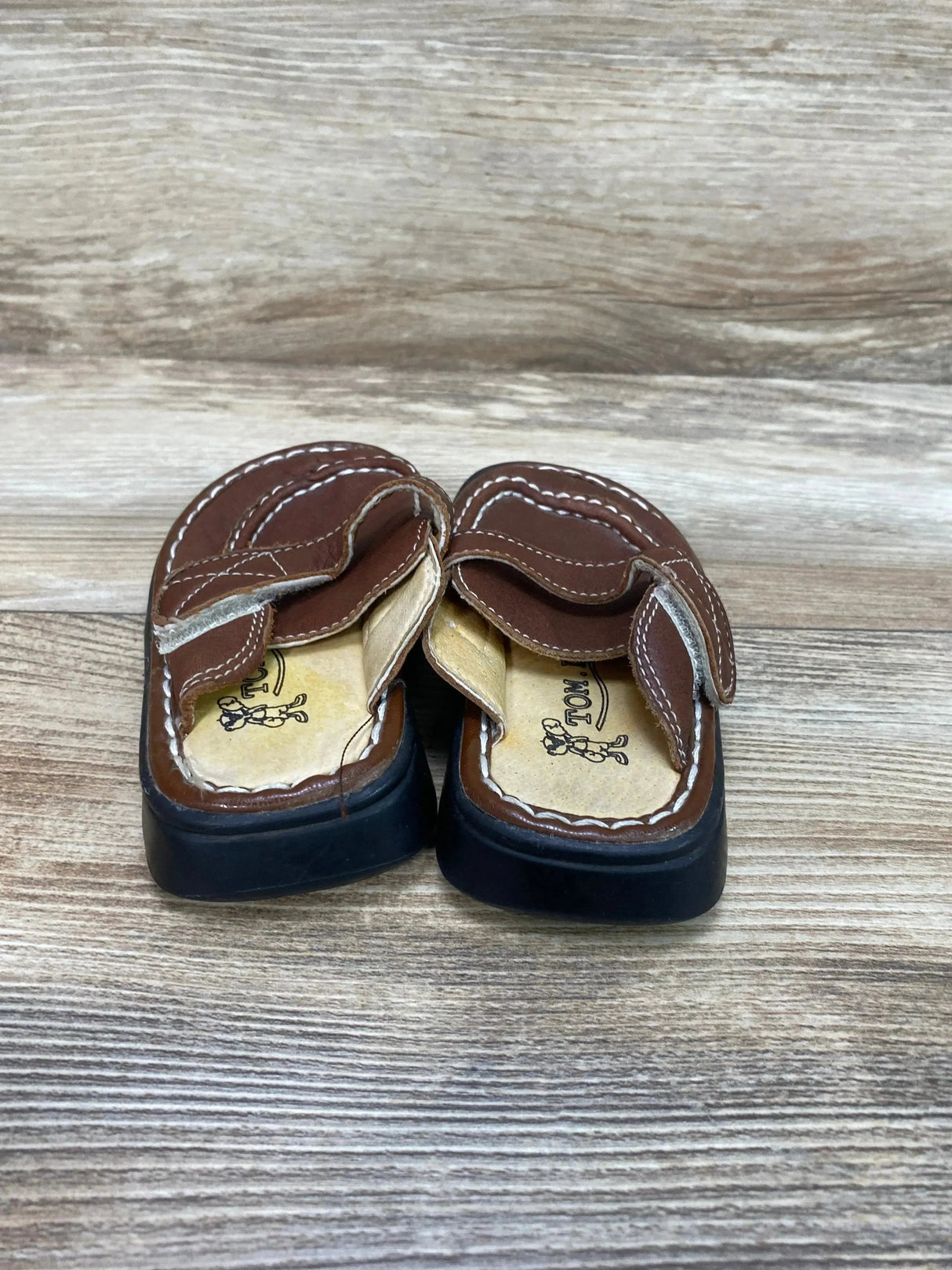 Front view of Tom Dog brown leather toddler clogs with Velcro strap and black sole on wood background, size 5/6C
