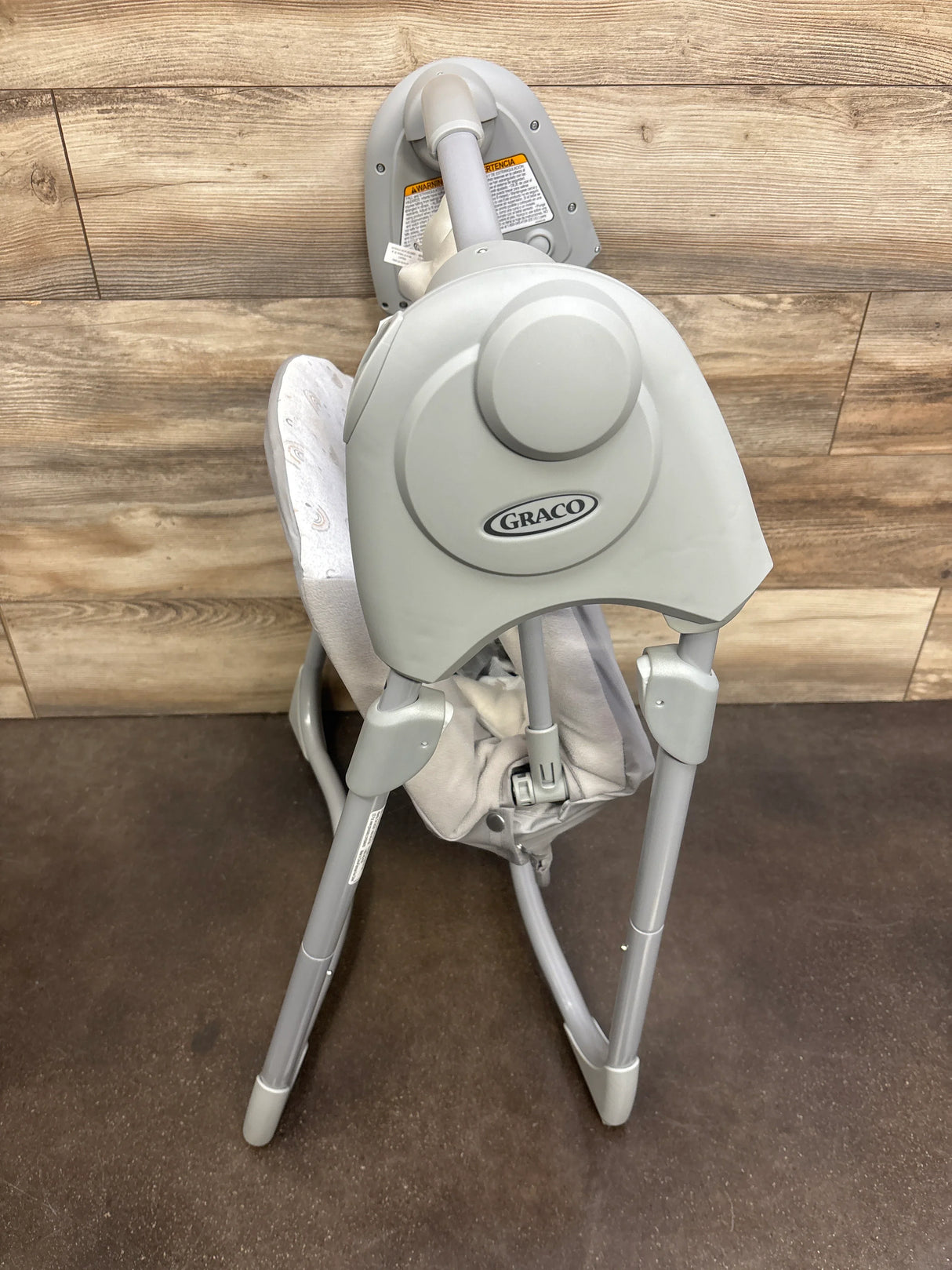 Rear three-quarter view of Graco Slim Spaces compact baby swing showcasing folded frame and Graco logo against wood-paneled wall and brown floor