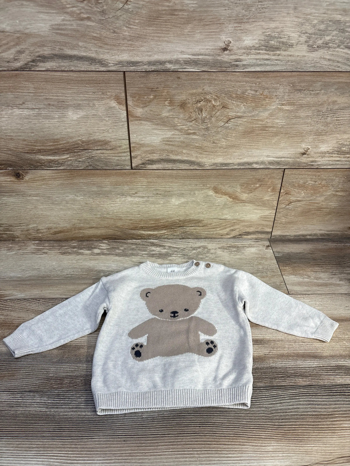Front view of H&M fine-knit jumper sweater in soft beige, showcasing a teddy bear design on a wood-look floor
