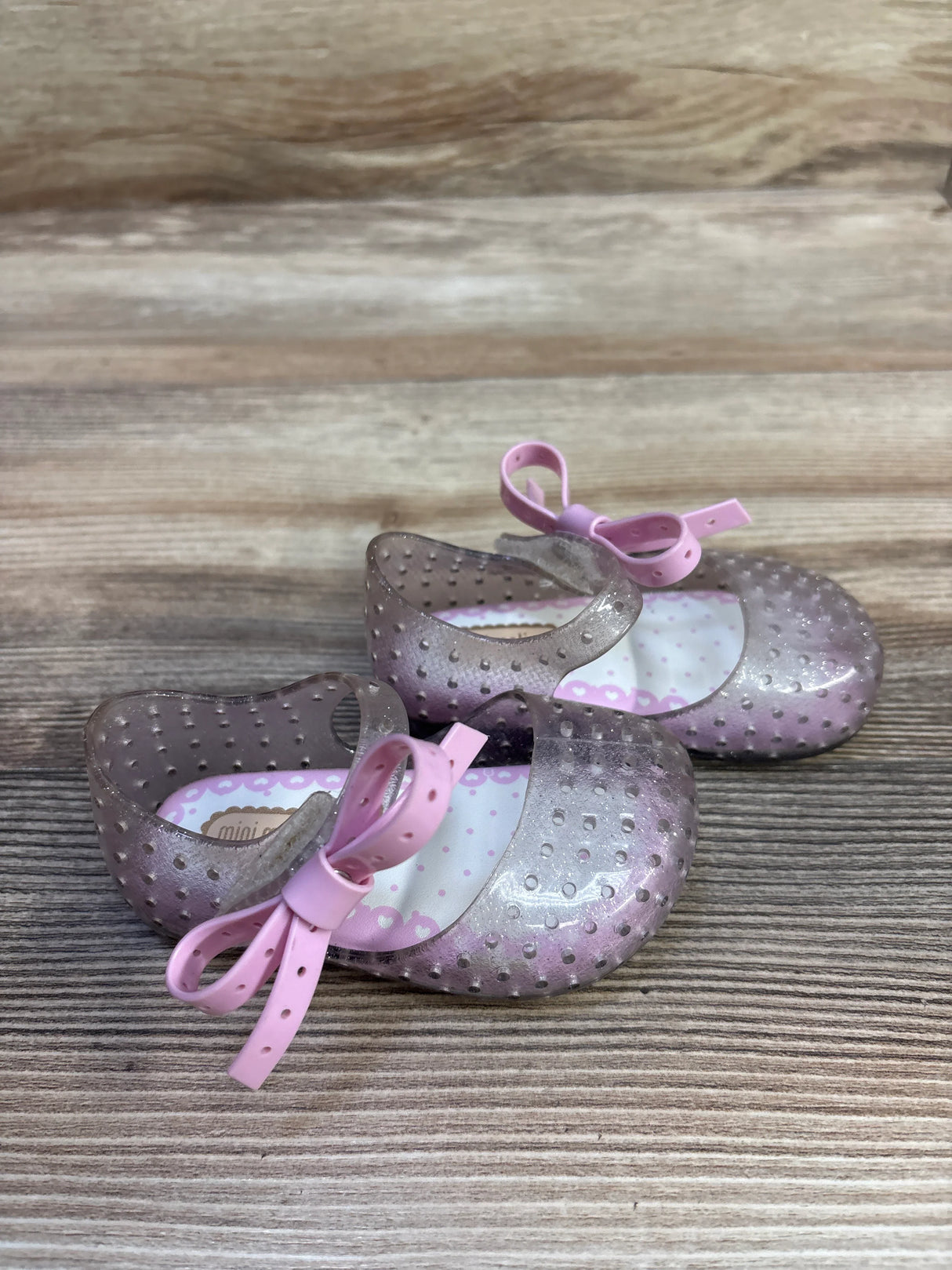 Angled front view of Mini Melissa Ultragirl jelly Mary Jane shoes with silver glitter, pink bow strap on wood background
