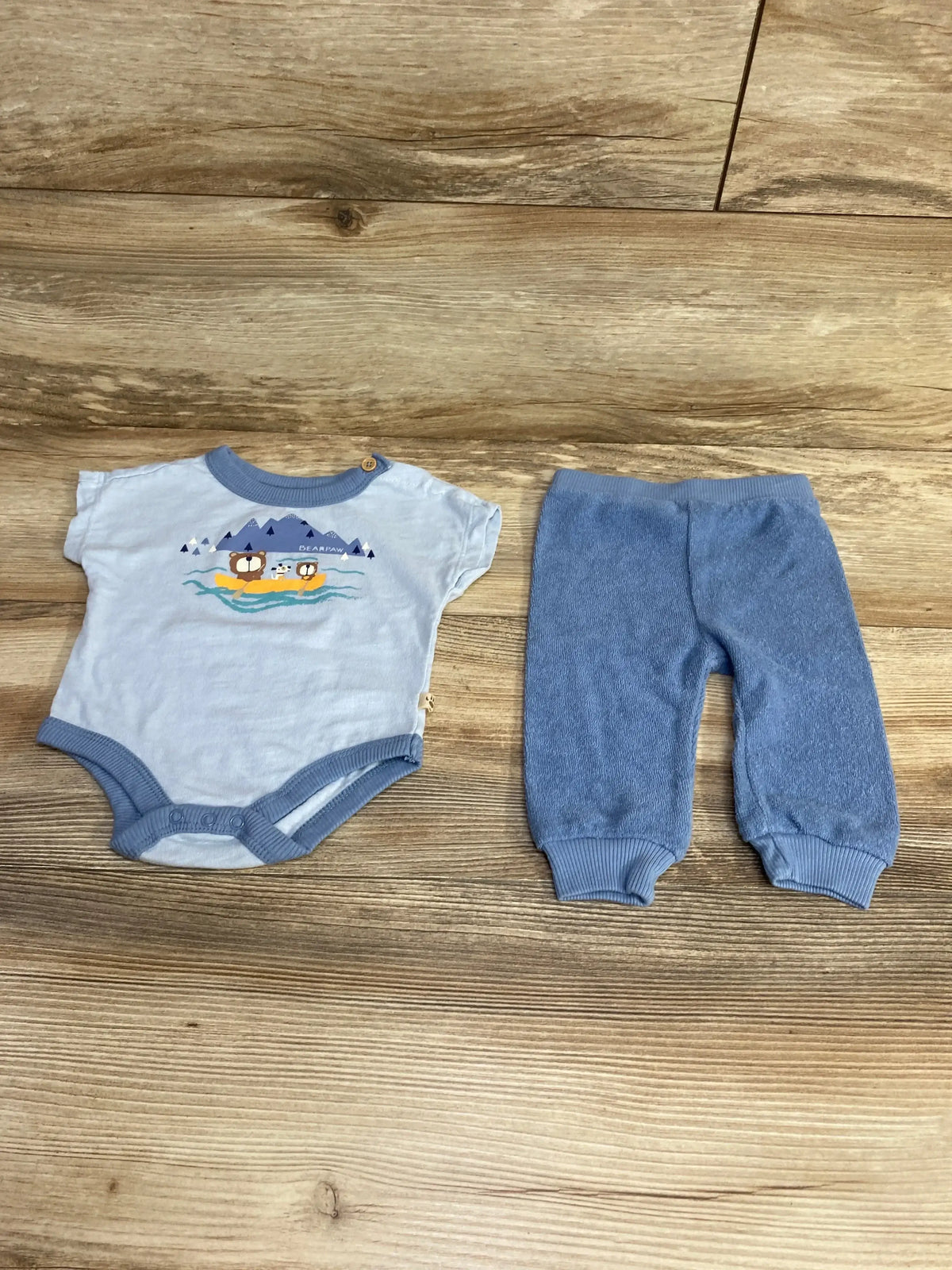 Flat lay of newborn blue Bearpaw bodysuit and pants showing bear graphic on bodysuit and ribbed cuffed ankles on wood floor