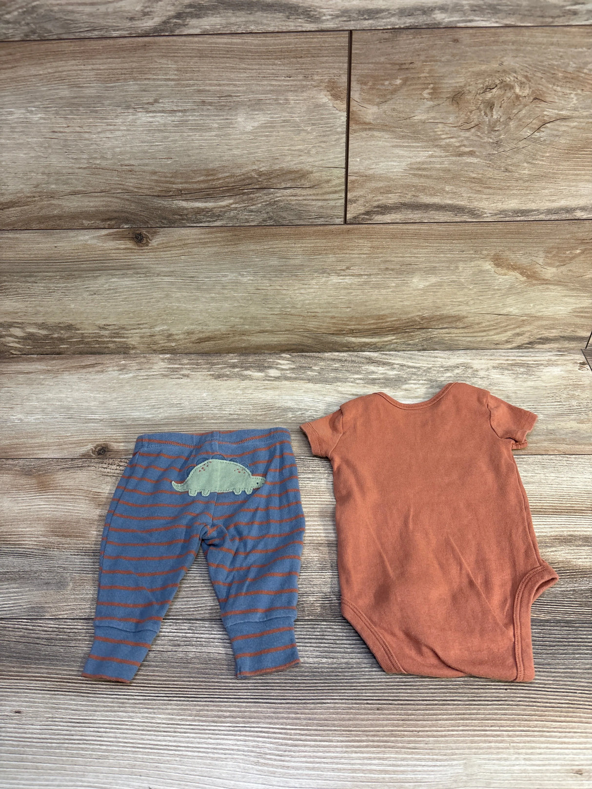 Flat lay of Carter’s 2-piece baby set showing rust-orange short-sleeve bodysuit and blue-striped pants with green dinosaur patch on wood background