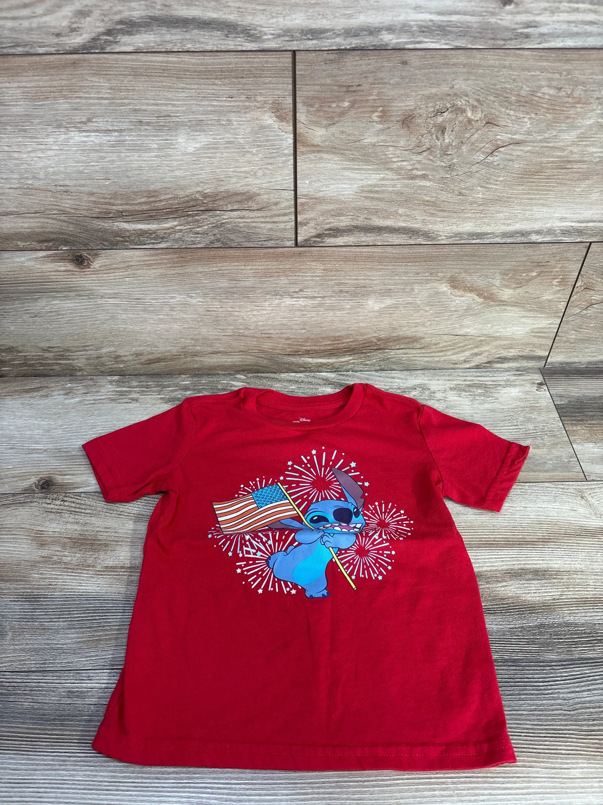 Flat-lay front view of red toddler T-shirt (5T) showing blue Stitch holding an American flag with fireworks print on a light wood floor
