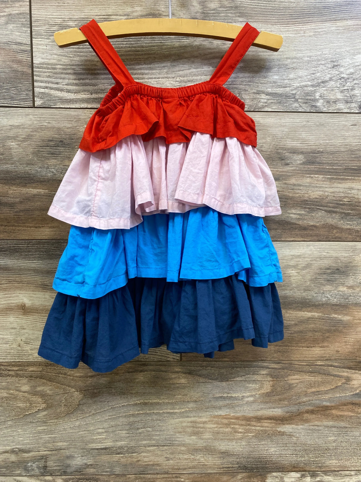 Front view of Tucker and Tate tiered toddler dress showcasing layered tiers in red, light pink, bright blue, and navy on wooden hanger against wood plank background