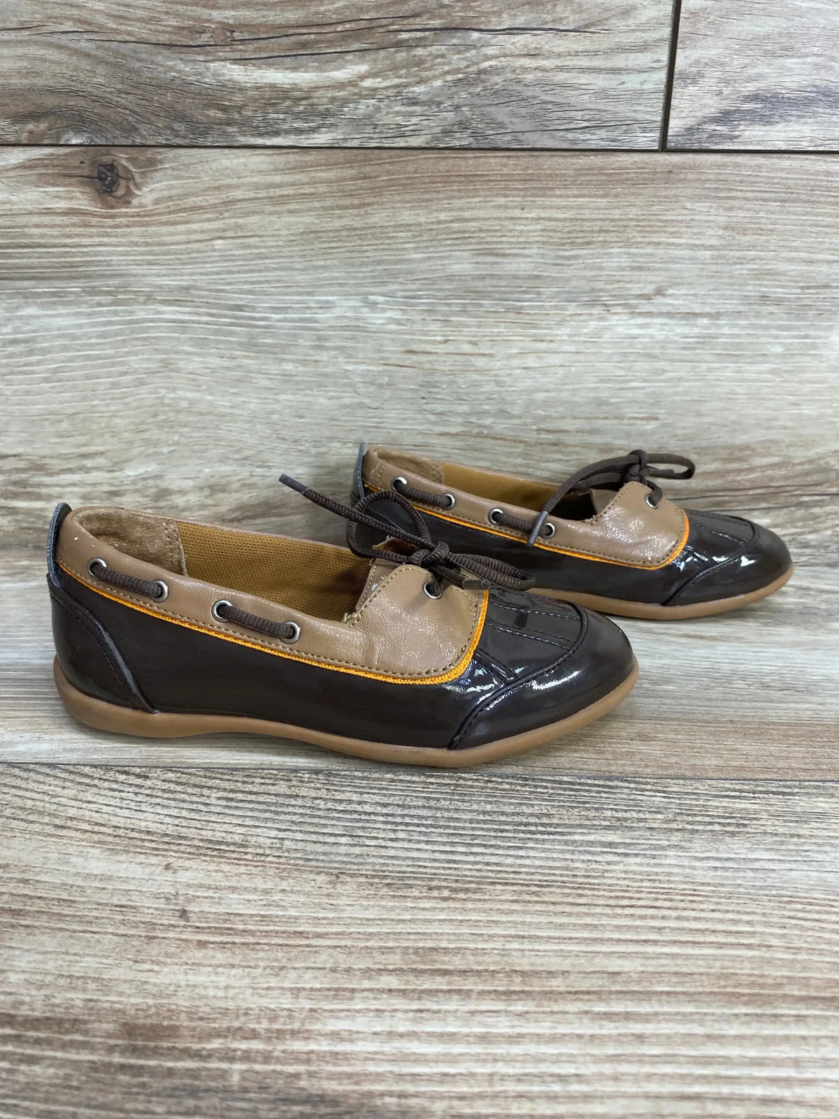 Side view of toddler brown patent boat shoes size 11C showcasing tan collar, orange piping, bow laces and gum sole on light wood background