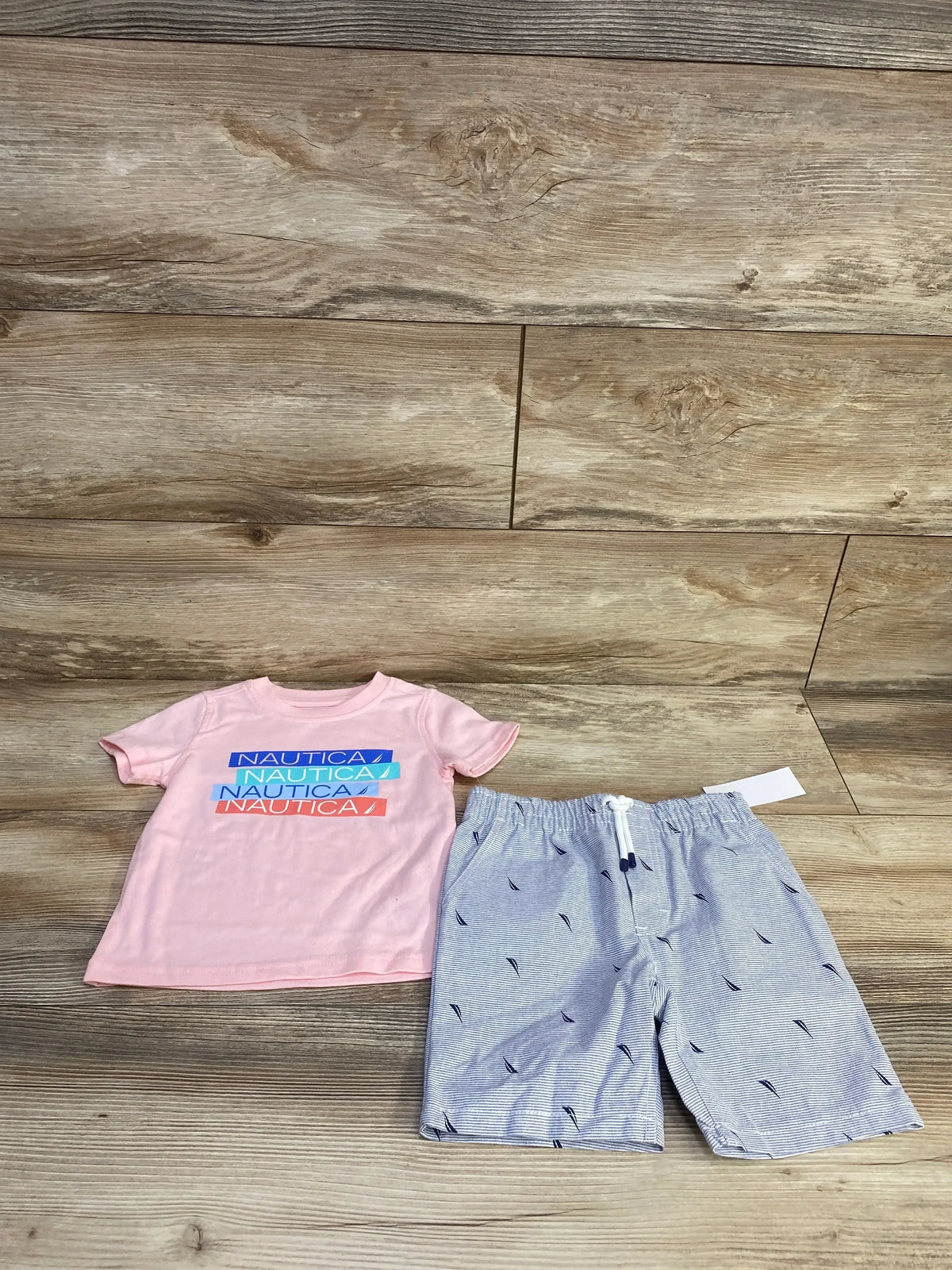 Flat lay of Nautica two-piece toddler set showing pink logo T-shirt and blue striped drawstring shorts on wood floor