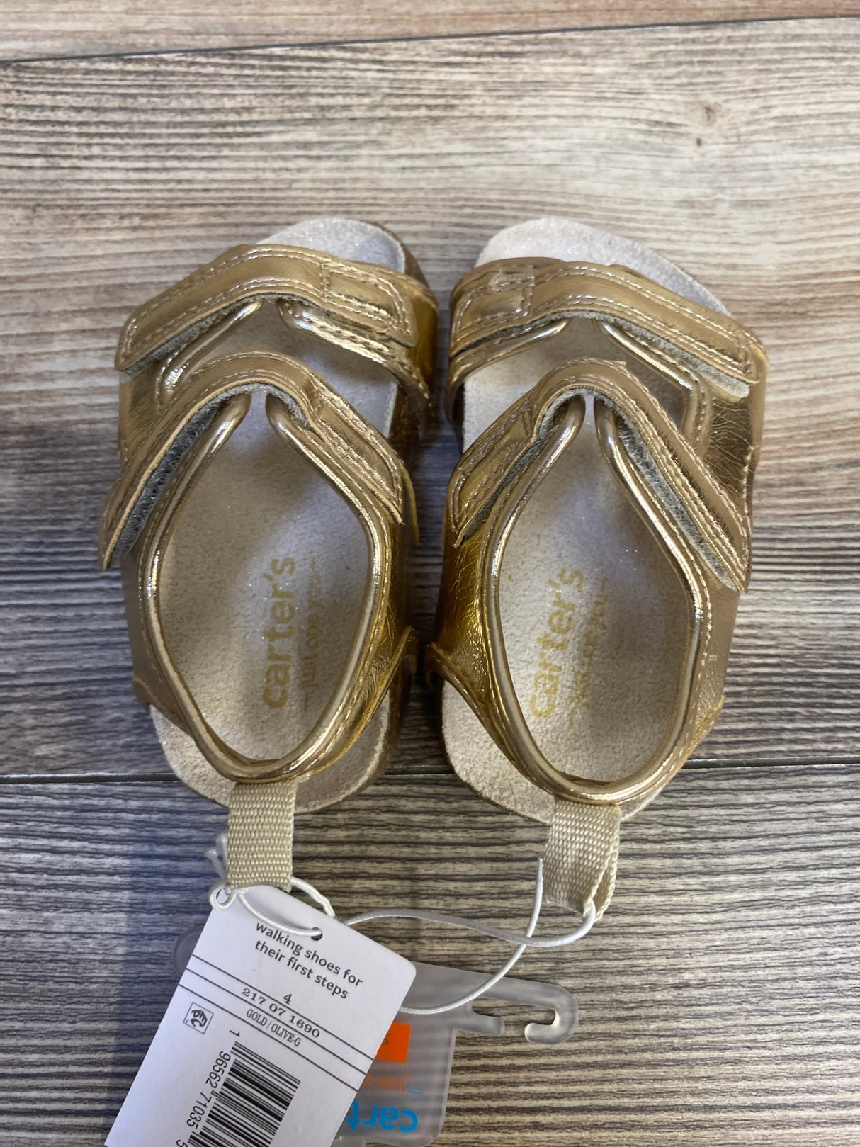 Top view of Just One You Olive First Walker sandals size 4C showcasing metallic gold finish, adjustable Velcro straps, and cork-effect insoles on a wood background